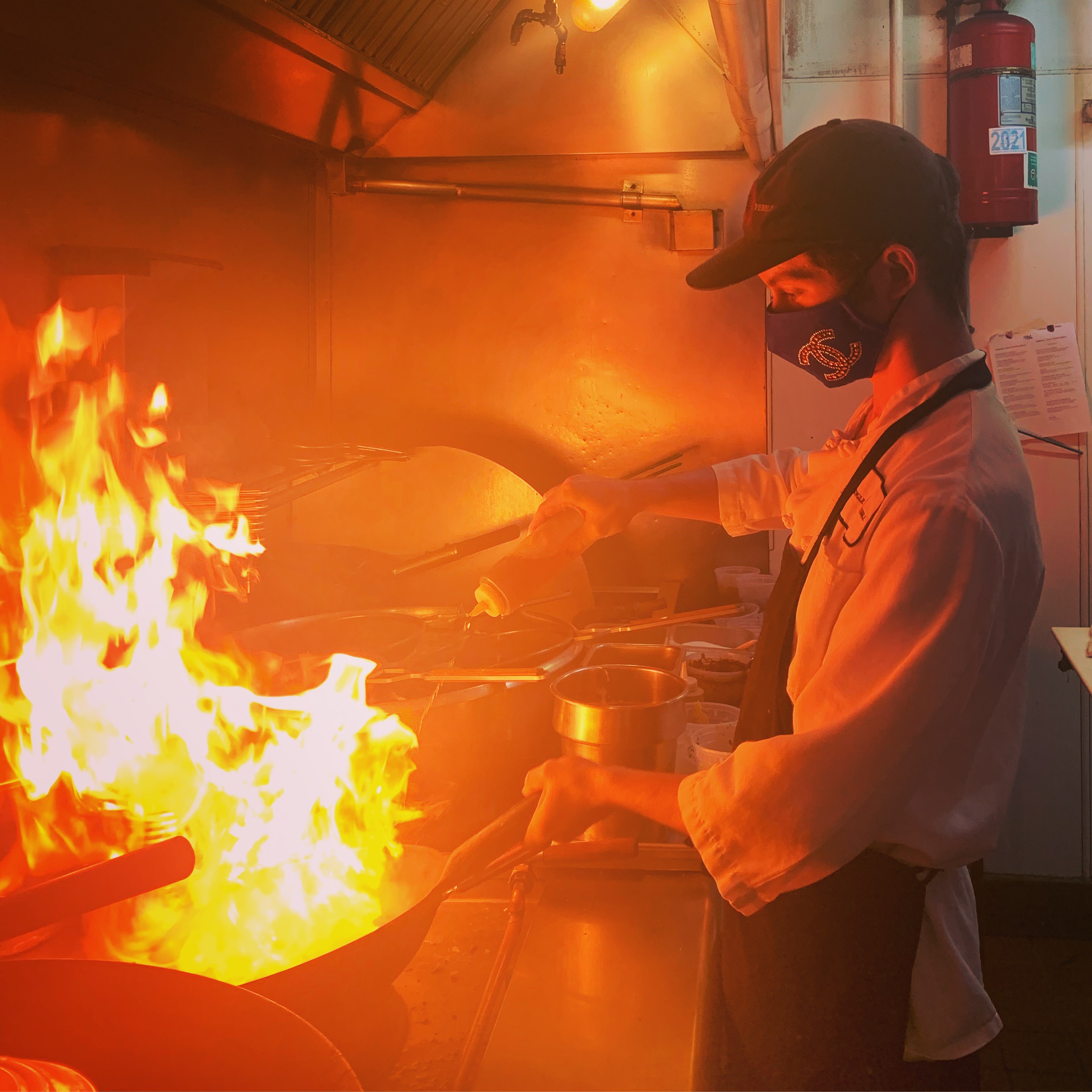 A man in a Chanel mask cooking in a kitchen. A wok has massive flames coming off it. 