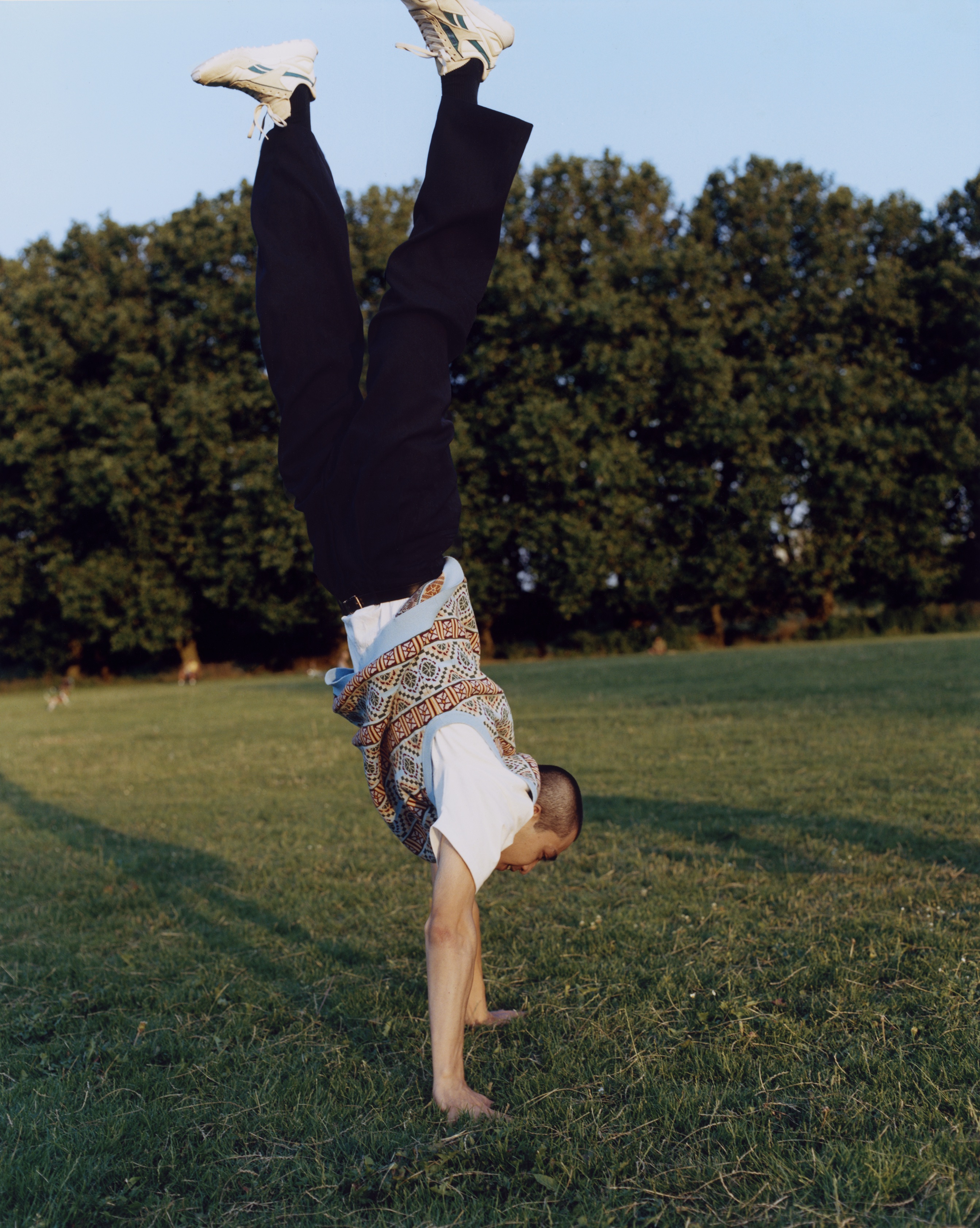 Makito does a handstand in a park. They wear white reeboks with black socks, black straight leg trousers, a white tee and a blue and red fair lisle print vest. 