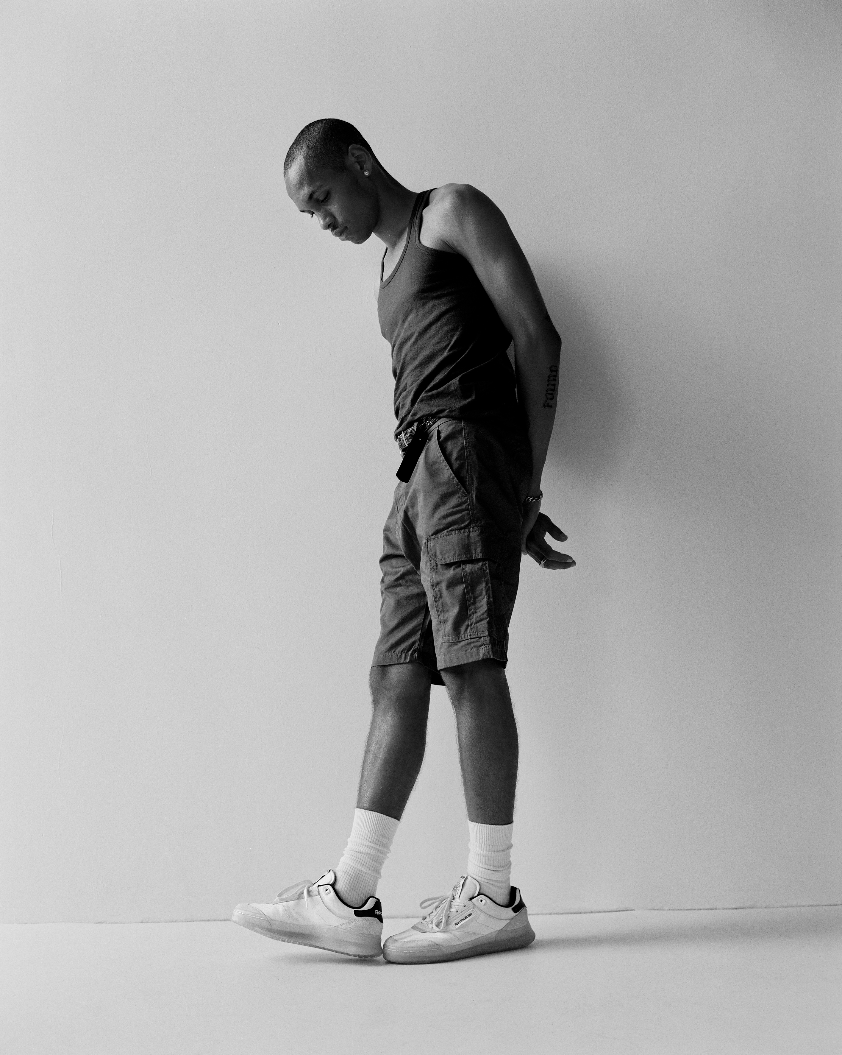 Tyrus stands in front of a white wall. They wear white reeboks with white socks, knee length utility shorts, a belt, and a grey vest.