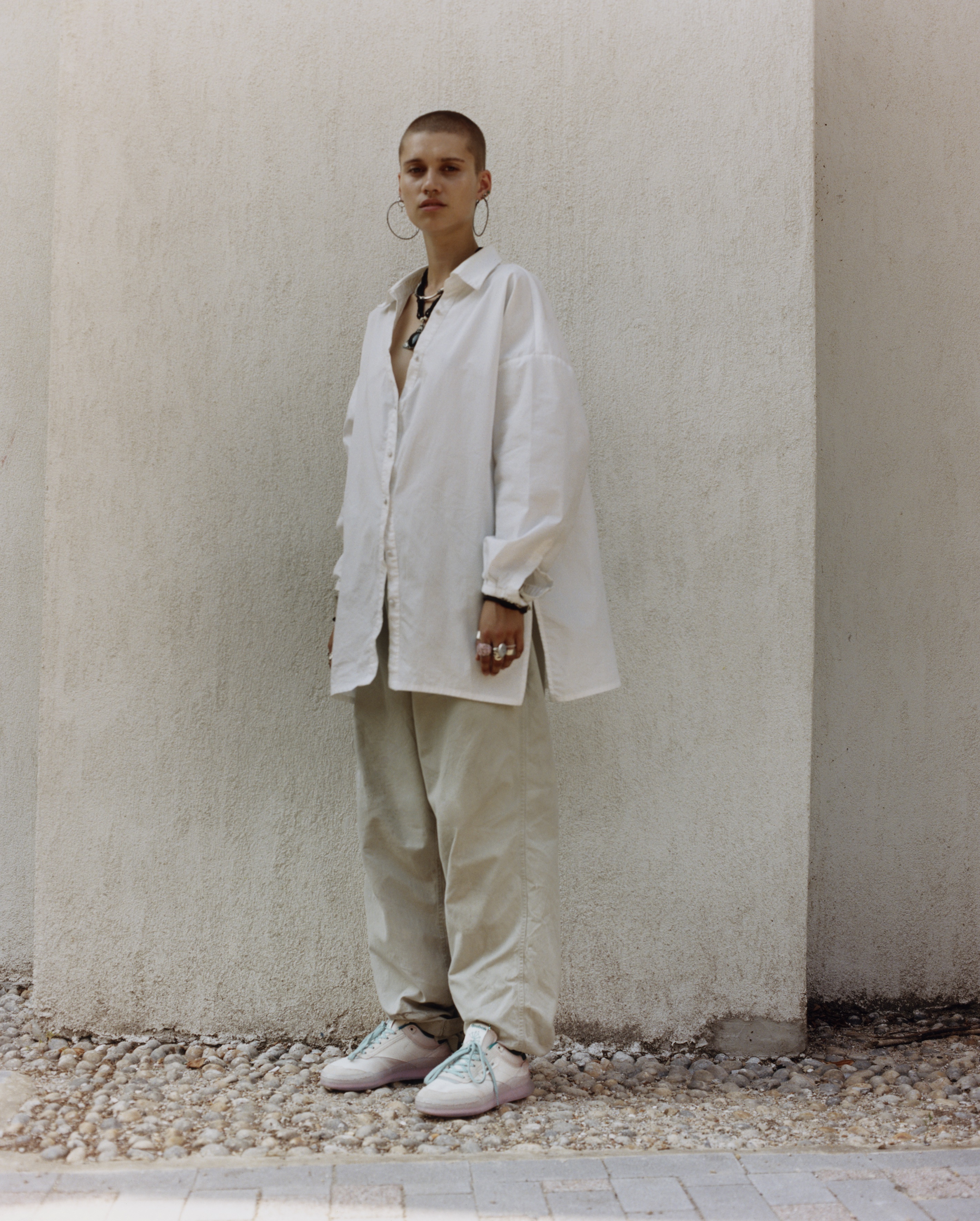 Isabelle stands on cobble stones in front of a white wall outside. They wear white reeboks with blue laces, sand coloured baggy trousers and a white oversized long sleeve shirt with lots of rings and a chunky necklace. 
