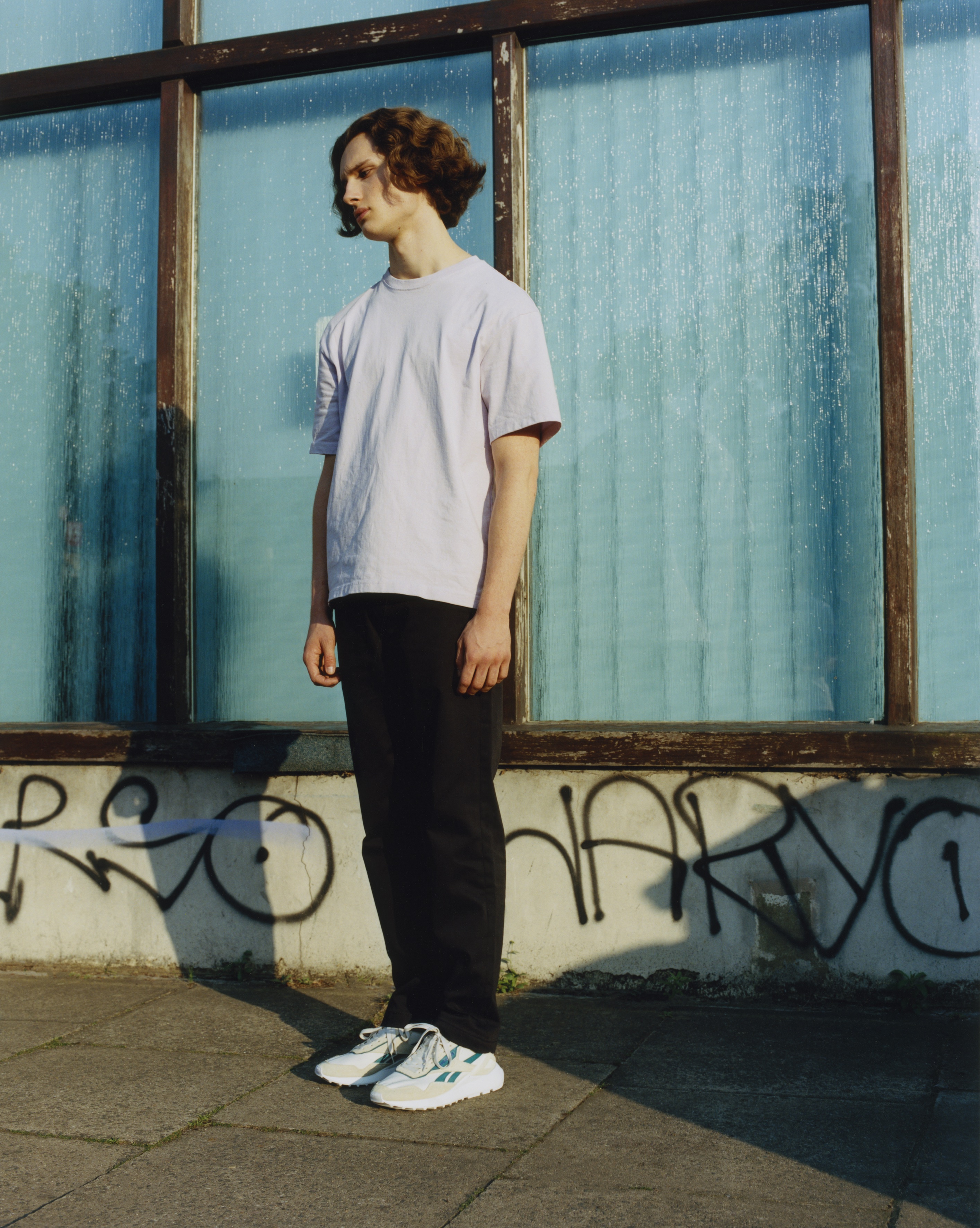 Laurie stands on the pavement next to windows. They wear white reeboks with blue detailing. Back trousers and a white t-shirt.
