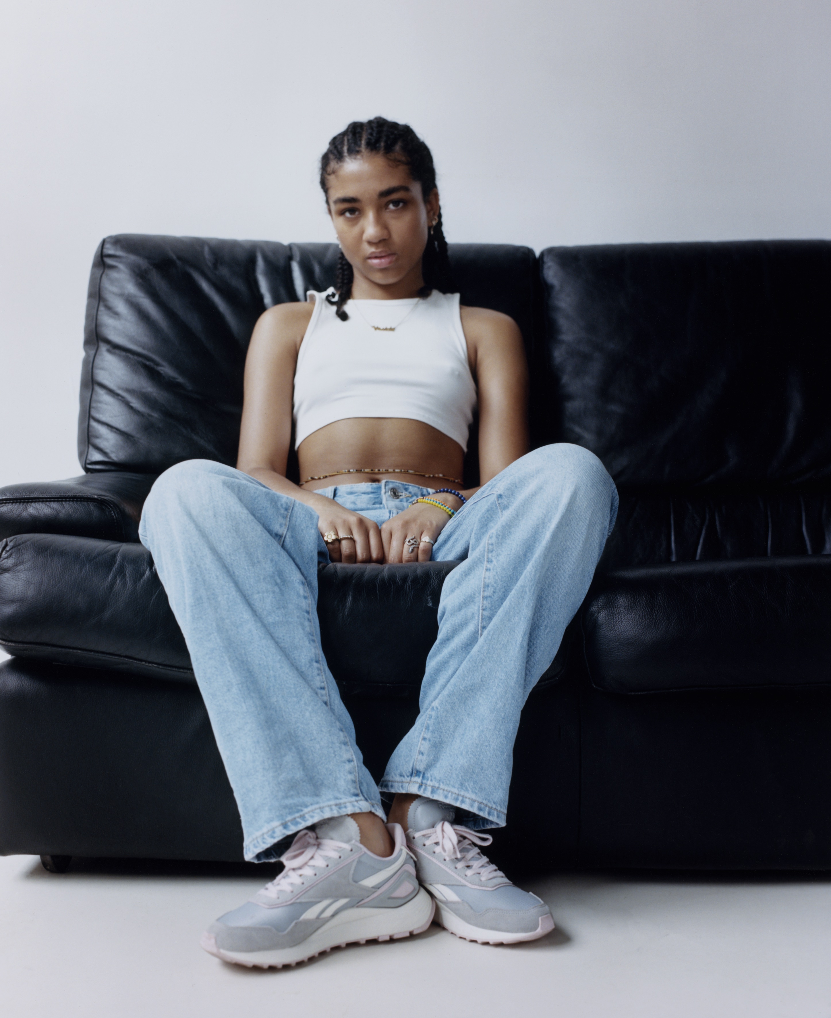 Betty sits on a black leather sofa in front of a grey wall. They have grey reeboks with no socks, light blue straight fit jeans, a cropped white vest with a gold statement necklace, beaded belt around her abdomen and lots of beaded bracelets and rings. 
