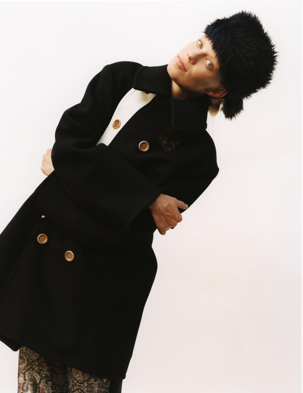 a woman in a double breasted coat leans to the side, a furry black hat on her head