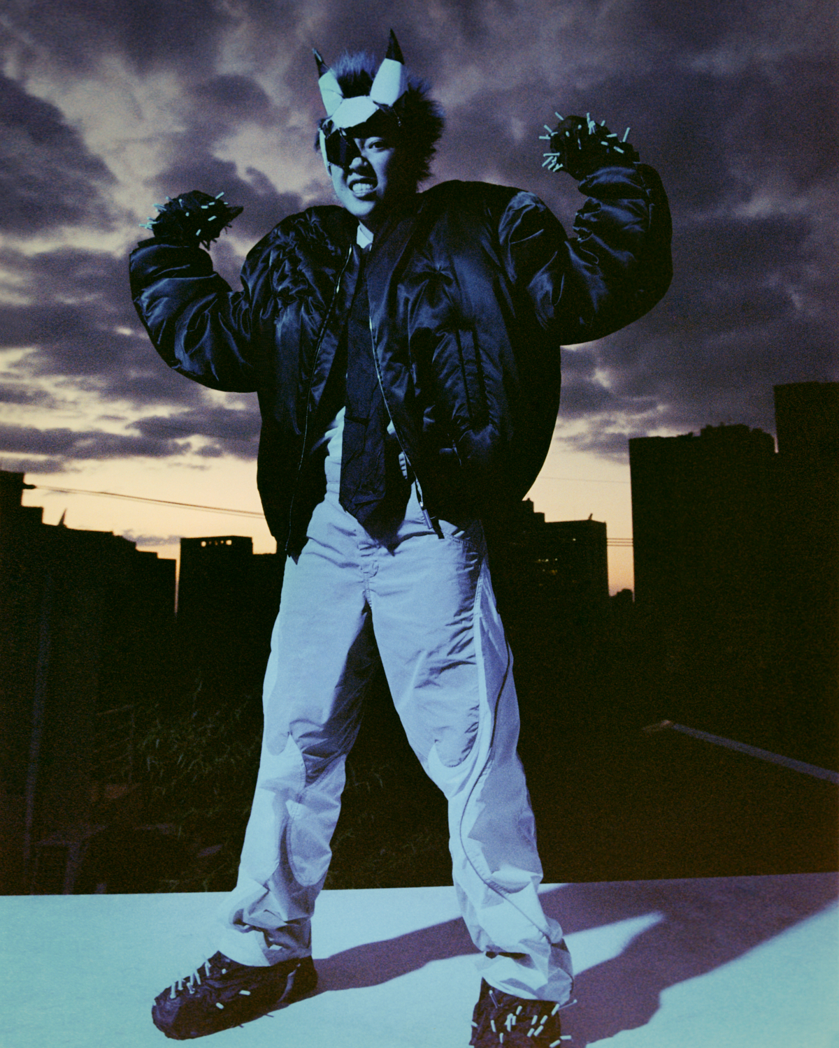 a photograph of a person wearing an eye patch, devil-like horns and a bomber jacket, standing on a nighttime rooftop flexing their muscles