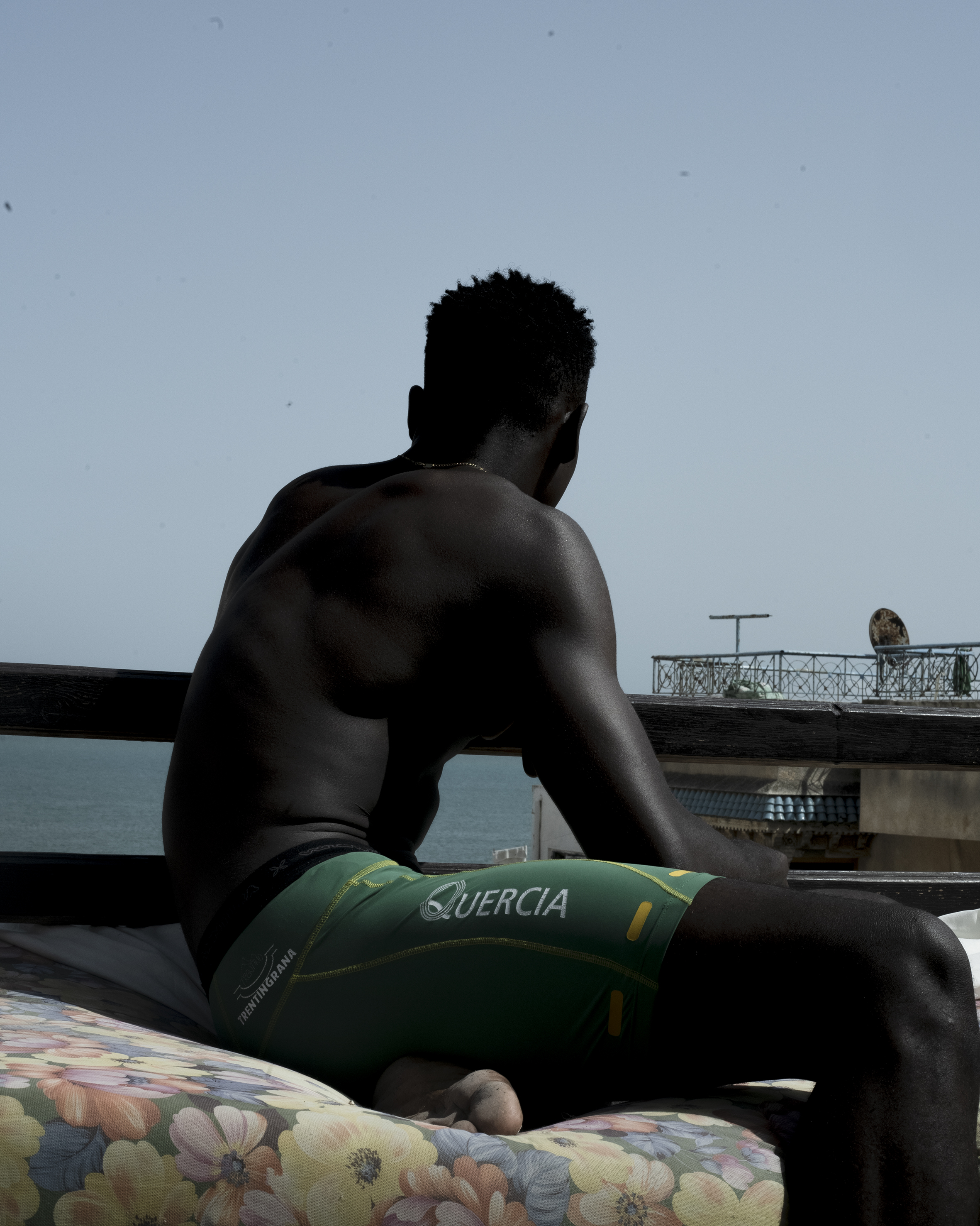 Man in shorts and shirtless sits on a floral cushion on a rooftop overlooking the ocean.