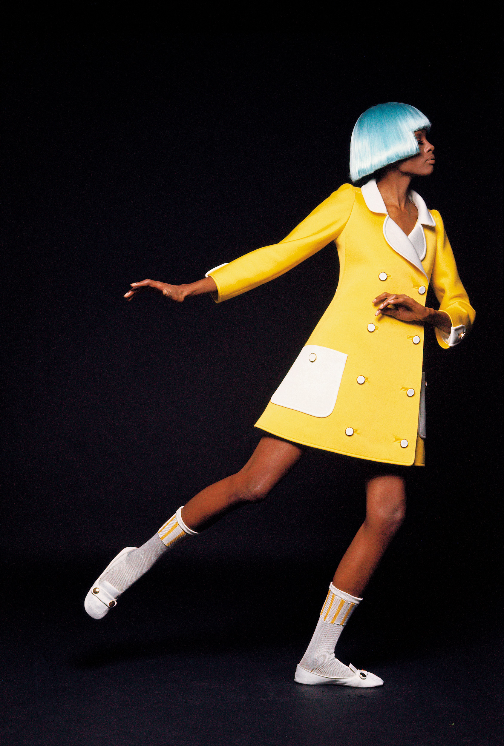 a woman in a blue wig and yellow coat posing in a studio