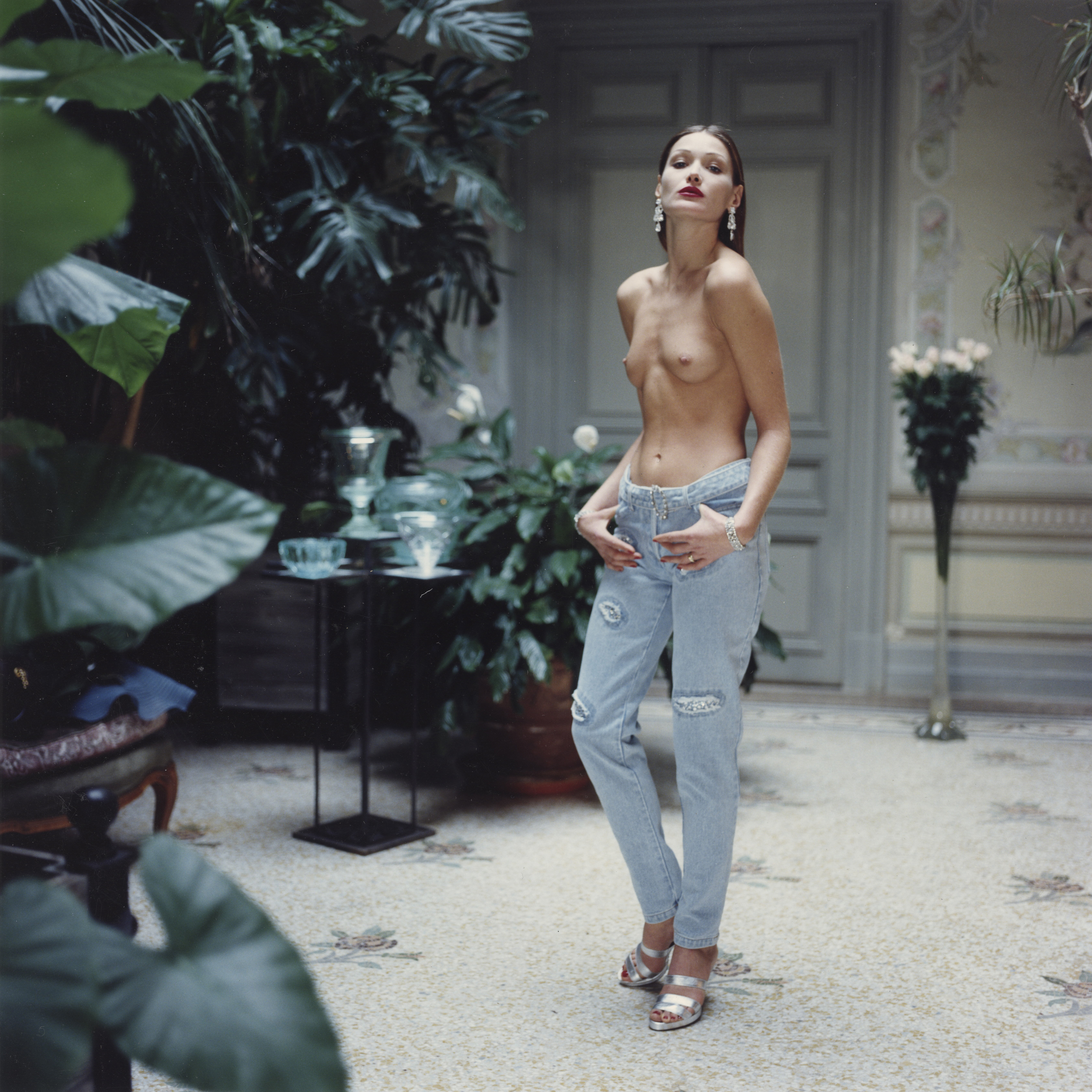 a woman wearing jeans, heels and no top with lots of plants around her