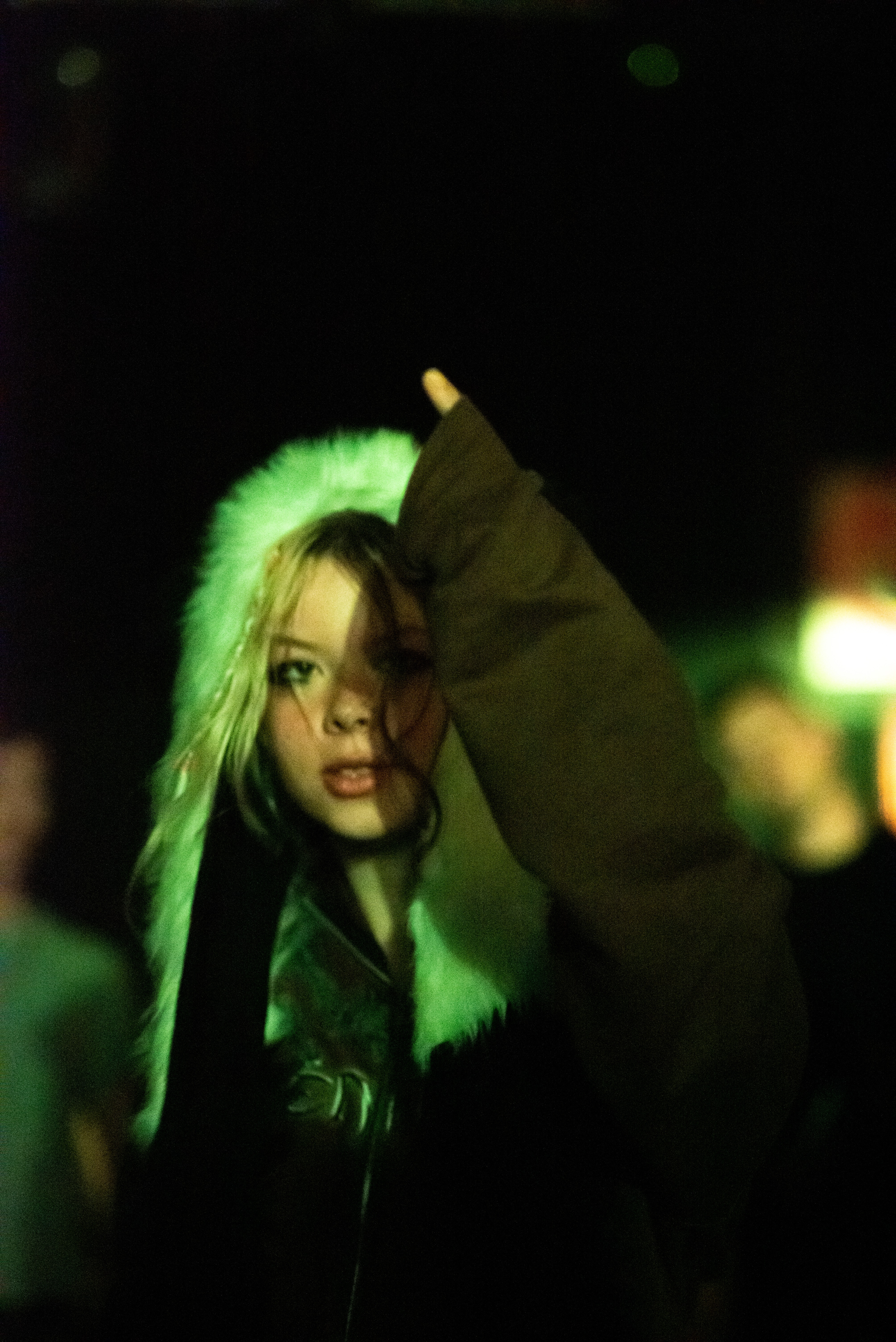 Girl in a furry hood and a dark coat puts her finger up standing in a crowd.