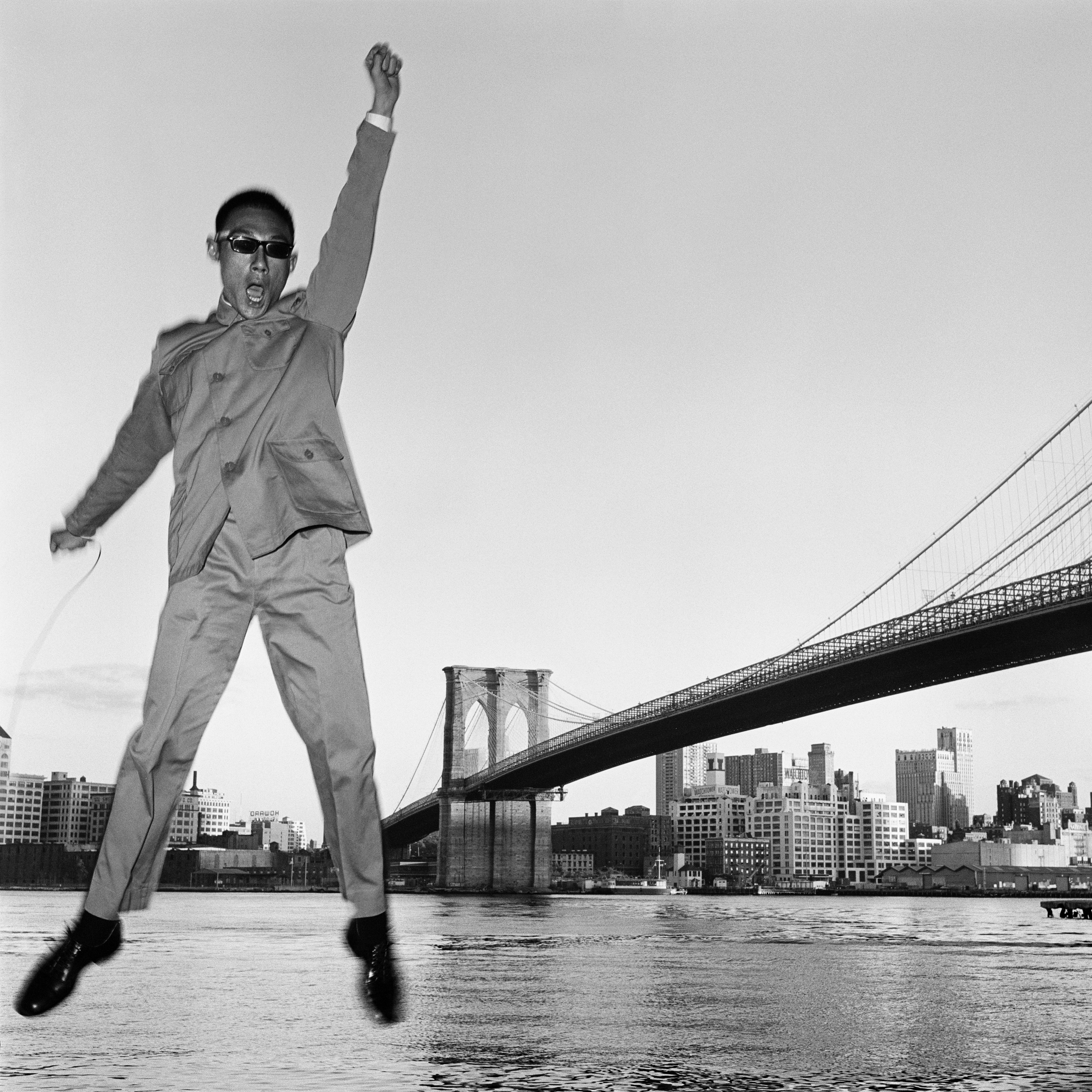 a man in a Zhongshan suit jumps in the air, taking a self portrait in front of a bridge in new york