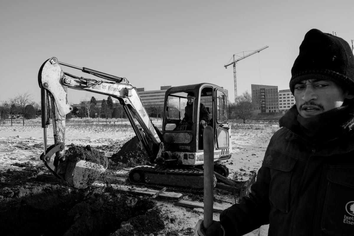 Jacques Vermeer, Patrick, cemetery - Black and white photo of a man in a beanie hat and jacket stood in front of a mechanical digger