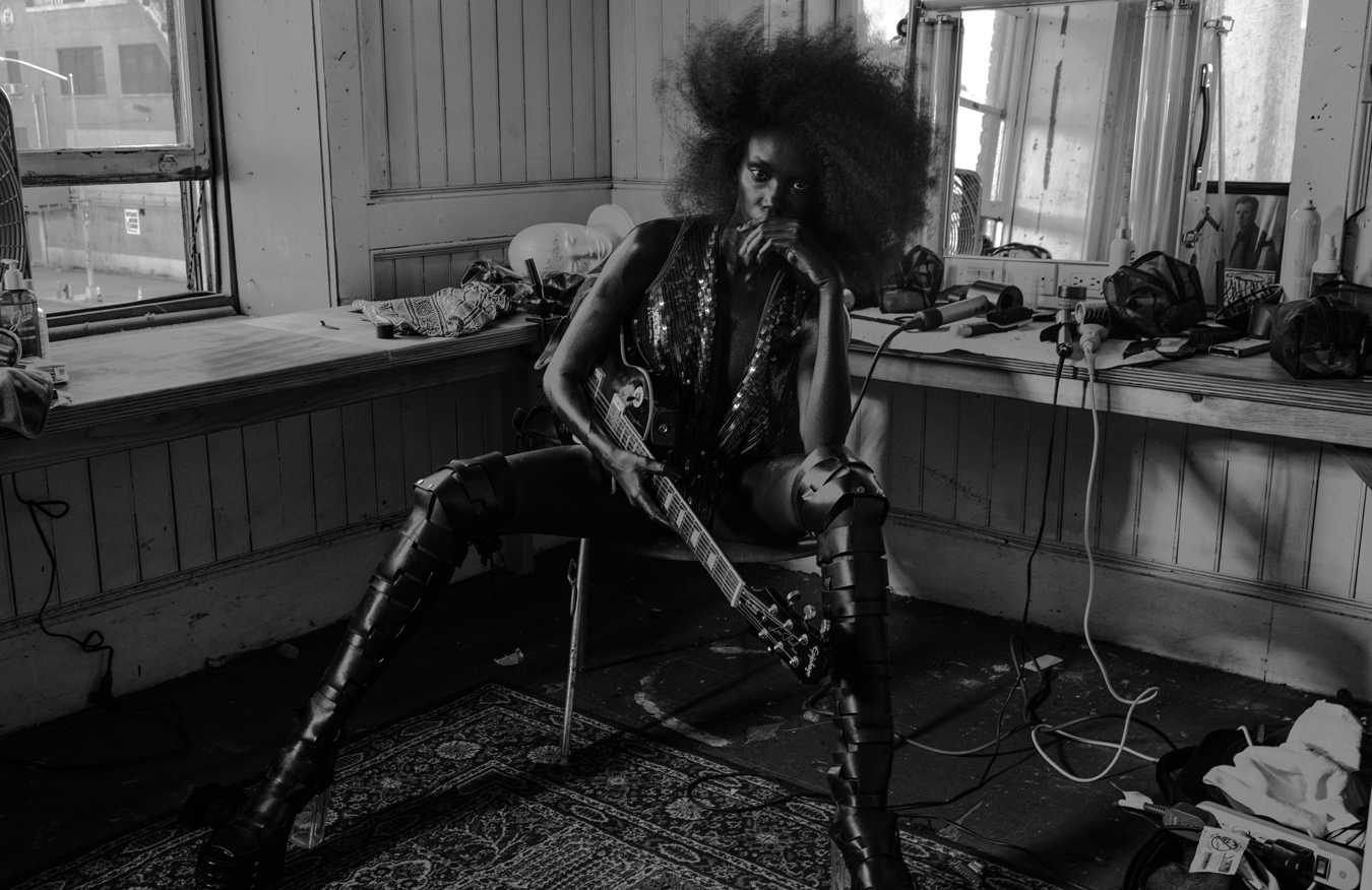 Anok Yai wears a sequin body suit, boots and a guitar next to a make up table and a mirror 