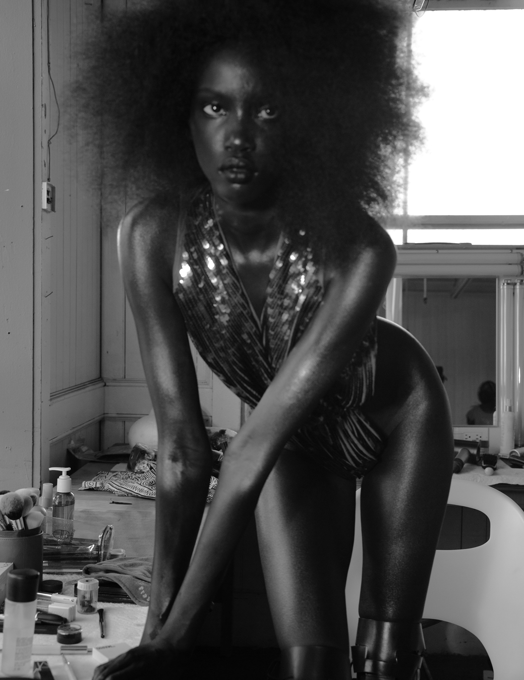 Anok Yai wears a sequin body suit next to a make up table and a mirror 