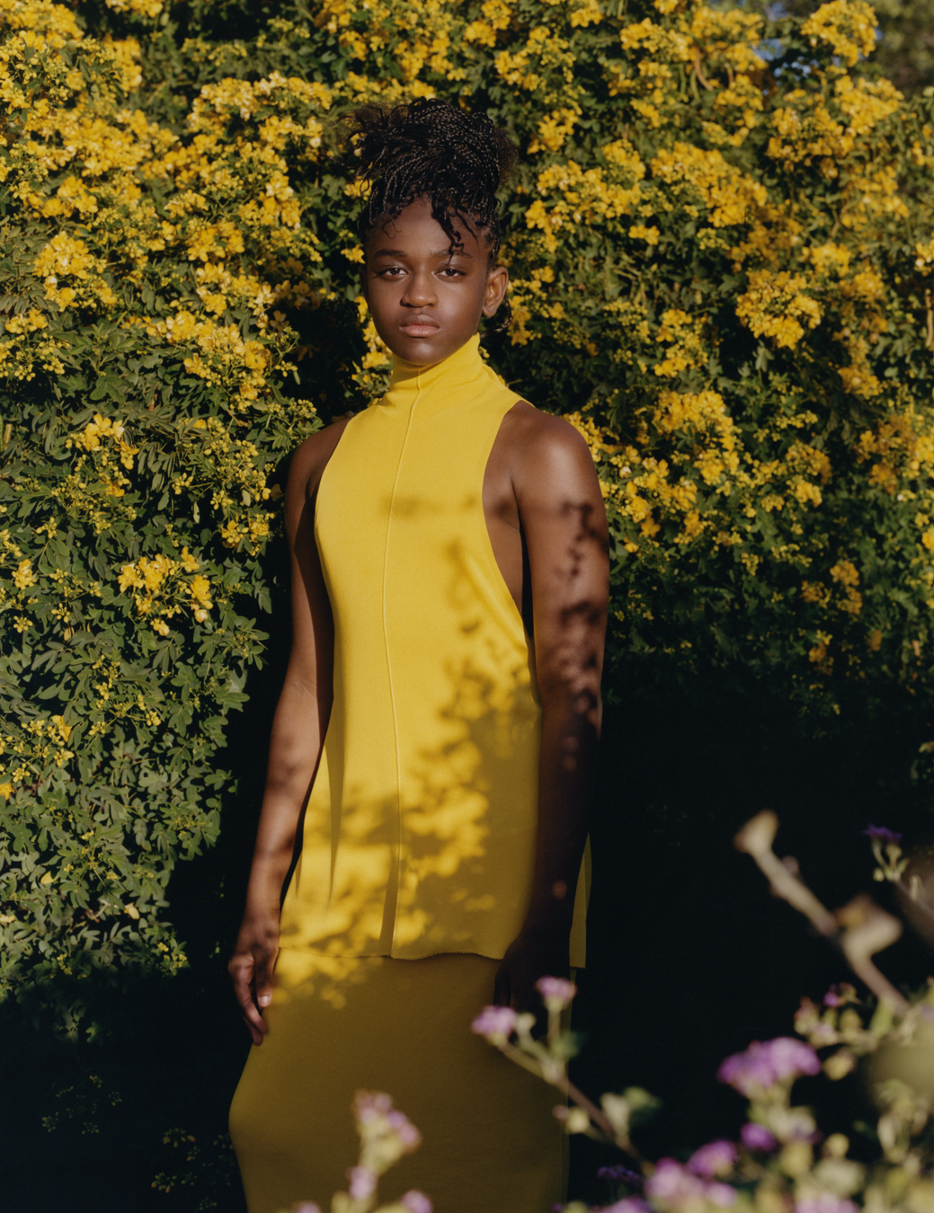Zaya wears a yellow tunic and yellow skirt standing in a bush with yellow flowers.