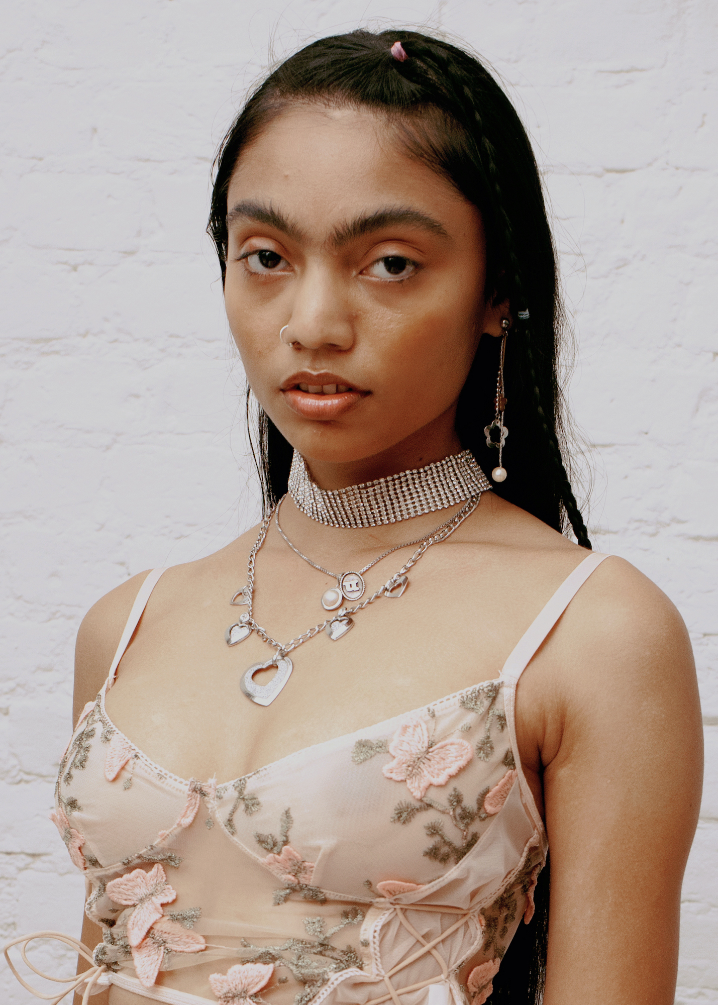 a girl in lots of silver jewellery and an embroidered bodice stands against a white brick wall