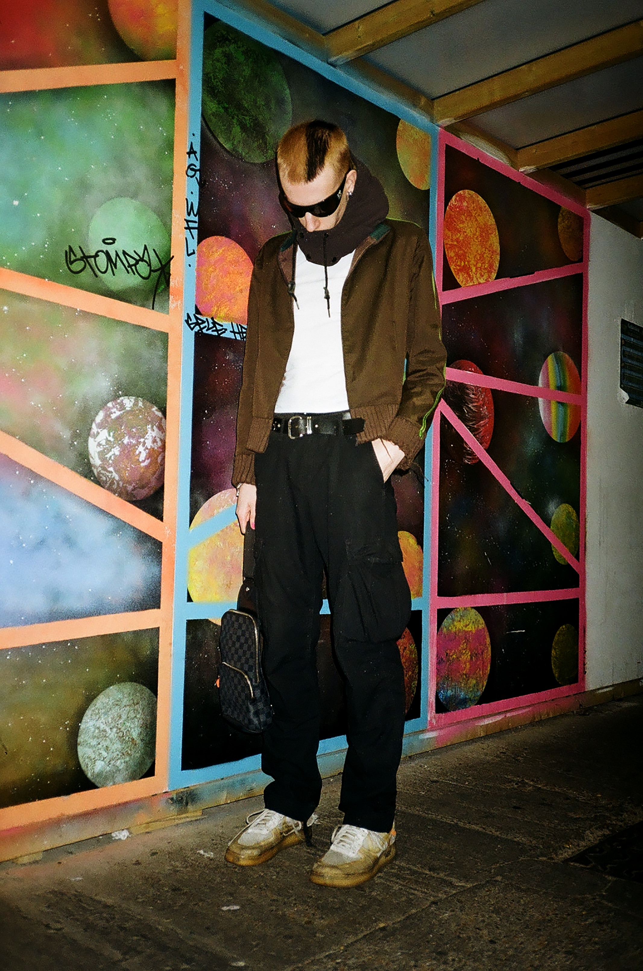 a guy with short half bleached, half dyed black hair stands wearing sunglasses against a mural of space
