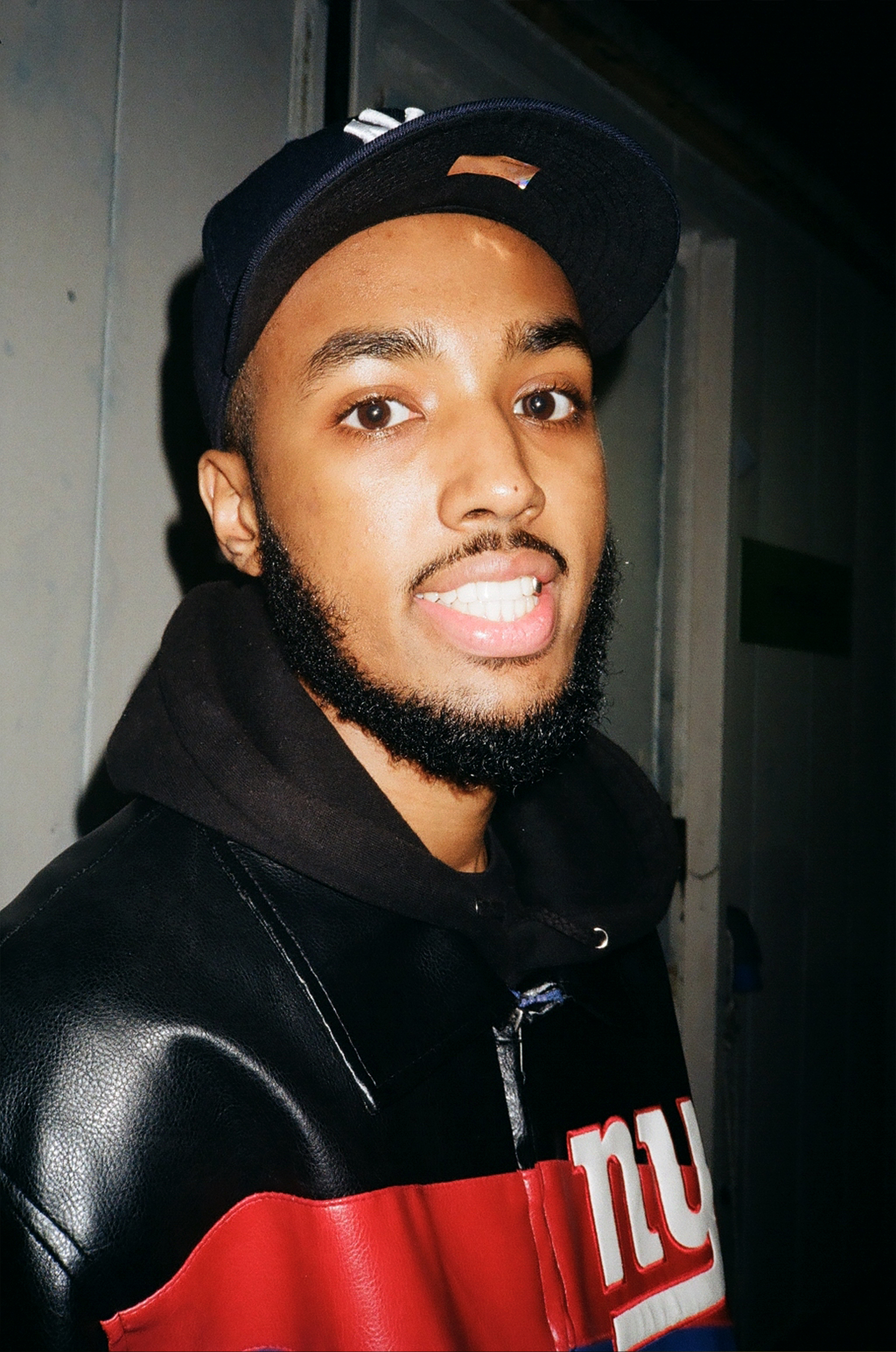 a young man with a beard, cap and leather jacket smiles to camera