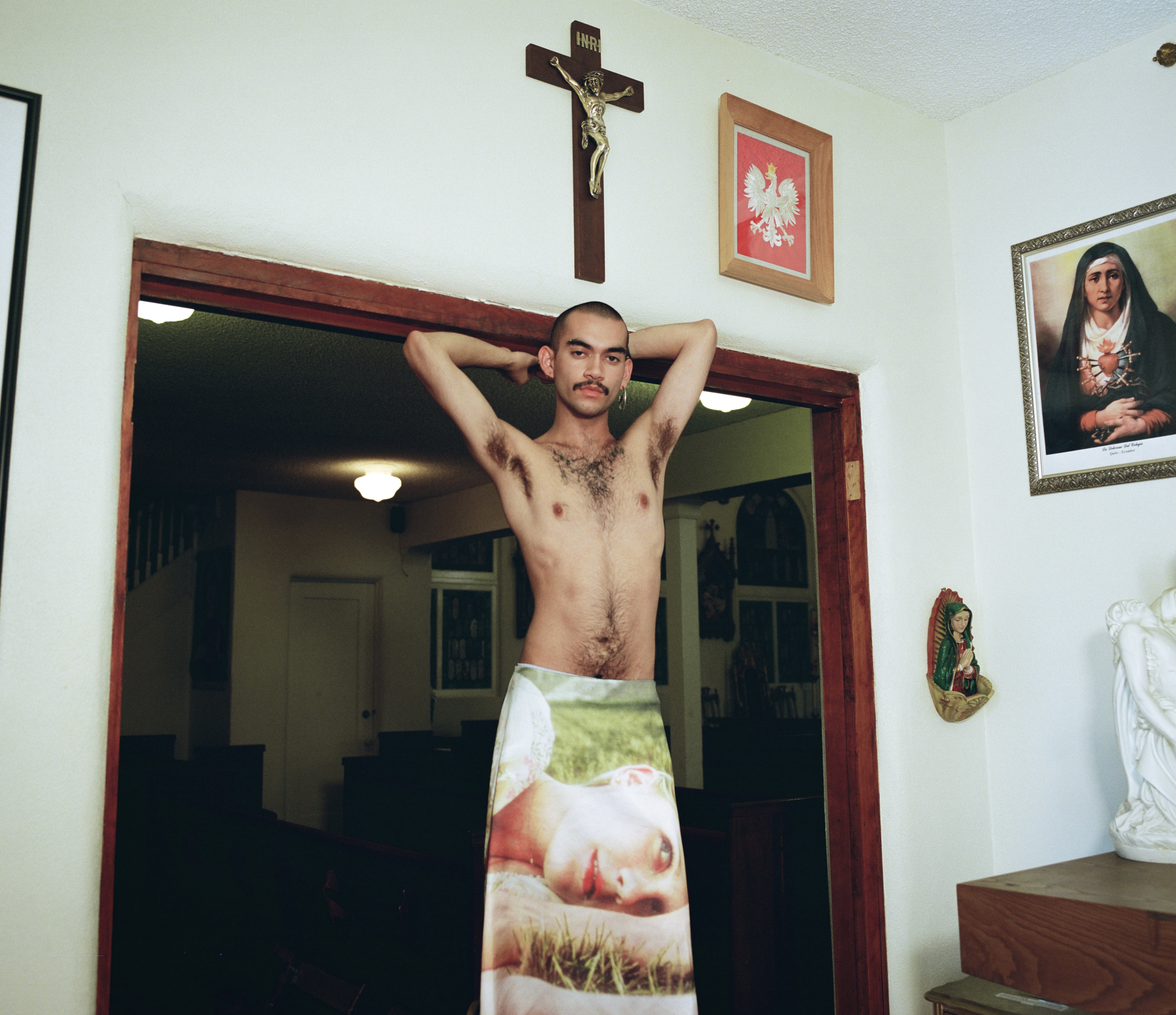 a topless male-presenting person stands in a room full of christian iconography wearing a heaven by marc jacobs skirt 