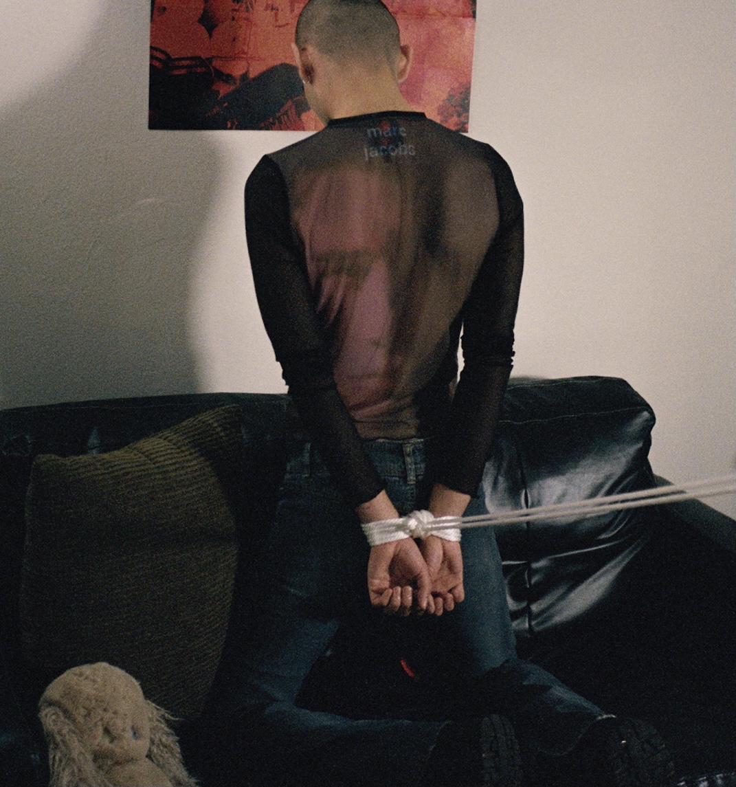a young male-presenting person with their wrists tied behind their back kneels on a leather sofa, facing away from the camera 