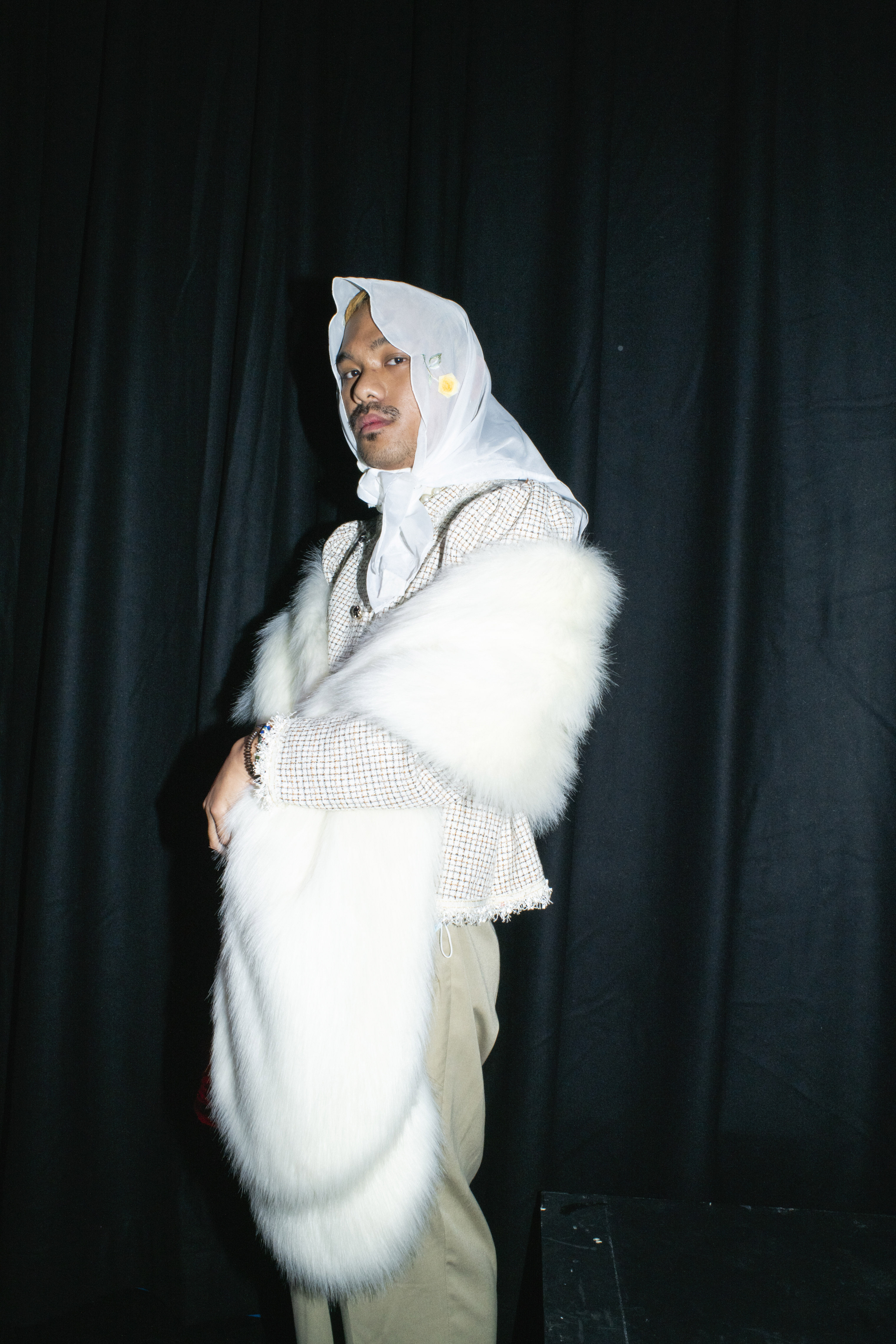 a man stands wearing a white faux fur stole and a chiffon headscarf over muted workwear trousers and a checked shirt