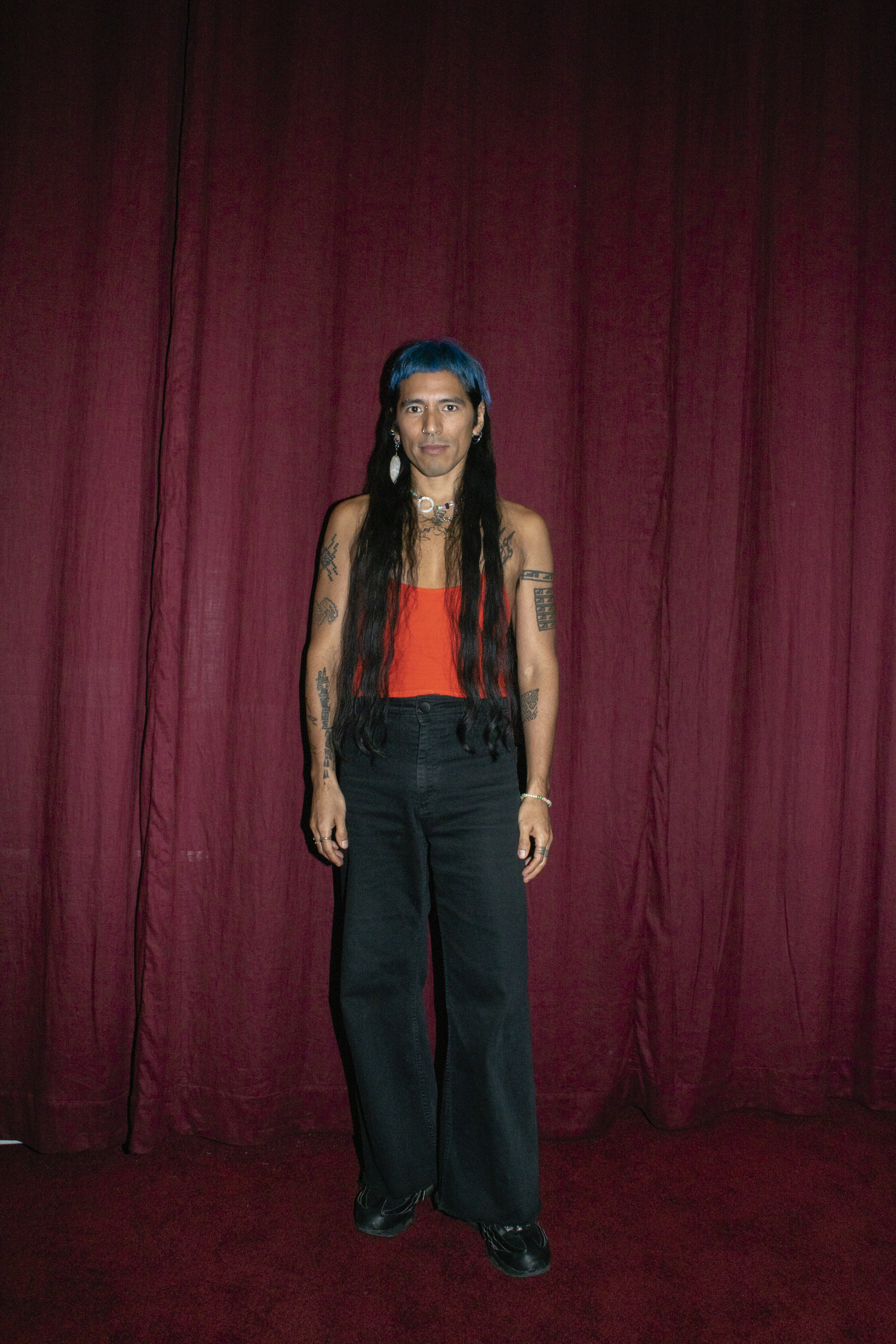 a person with tattoos and hip-length dark hair dyed bright blue at the roots stands in front of a red curtain