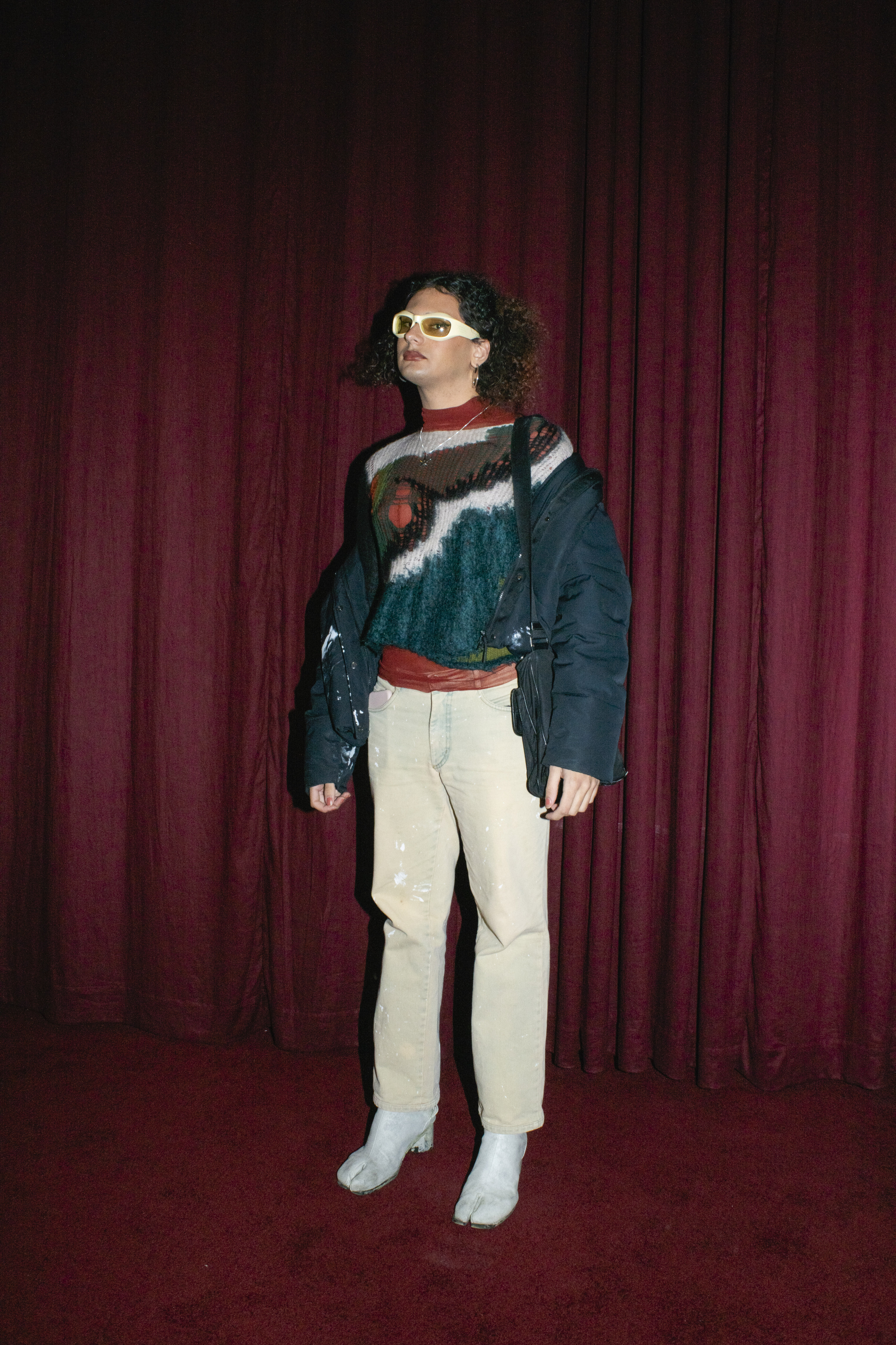 a person with a colourful knit jumper, pale jeans and tabi boots stands in front of a red curtain 