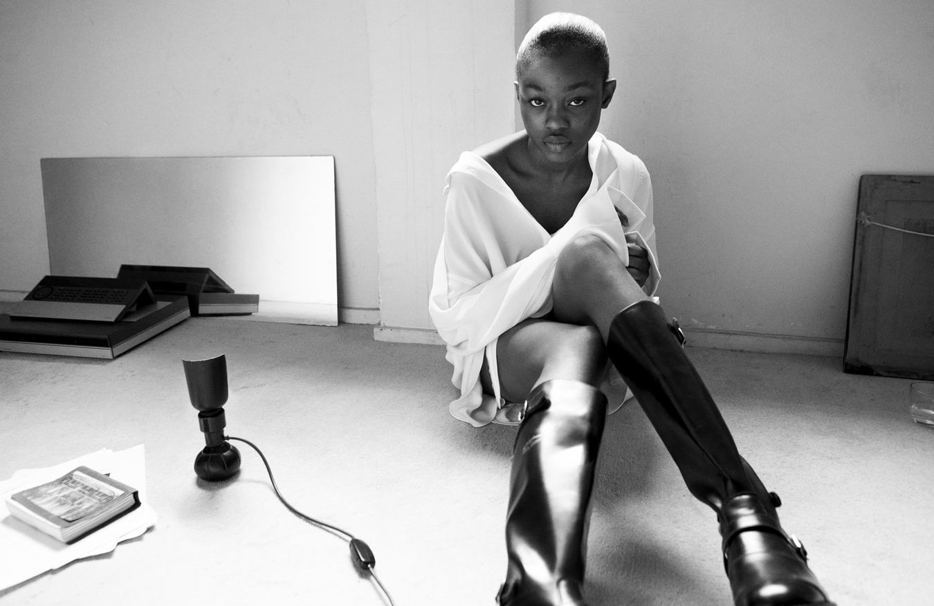 Model sits on the floor next to a mirror, lamp and book wearing a white shirt and black boots.