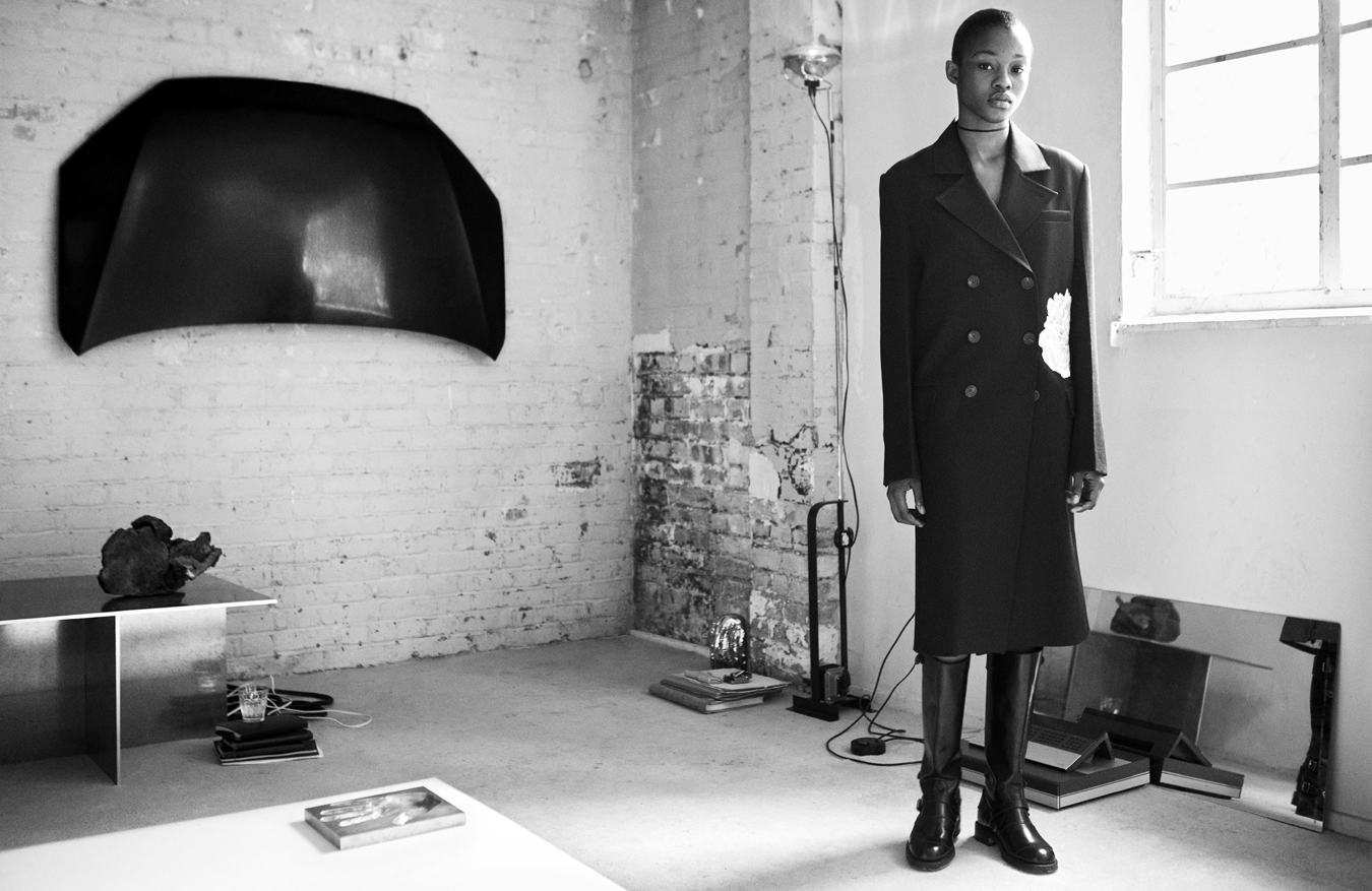 Model stands in a room with exposed brick wearing a black trench coat and black boots. 