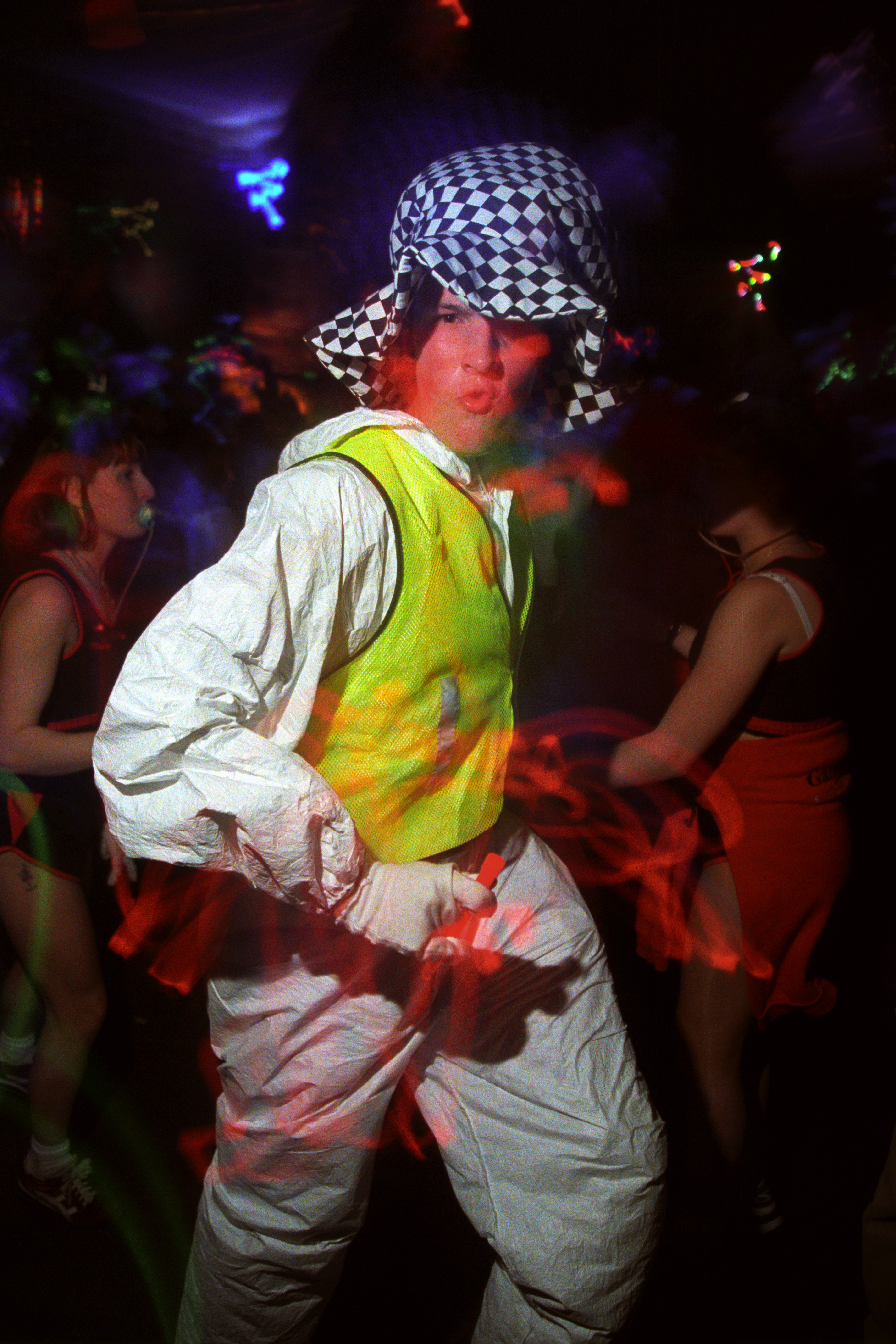 a man in a white boiler suit, high vis vest and checkered hat dances at a rave in the 90s 