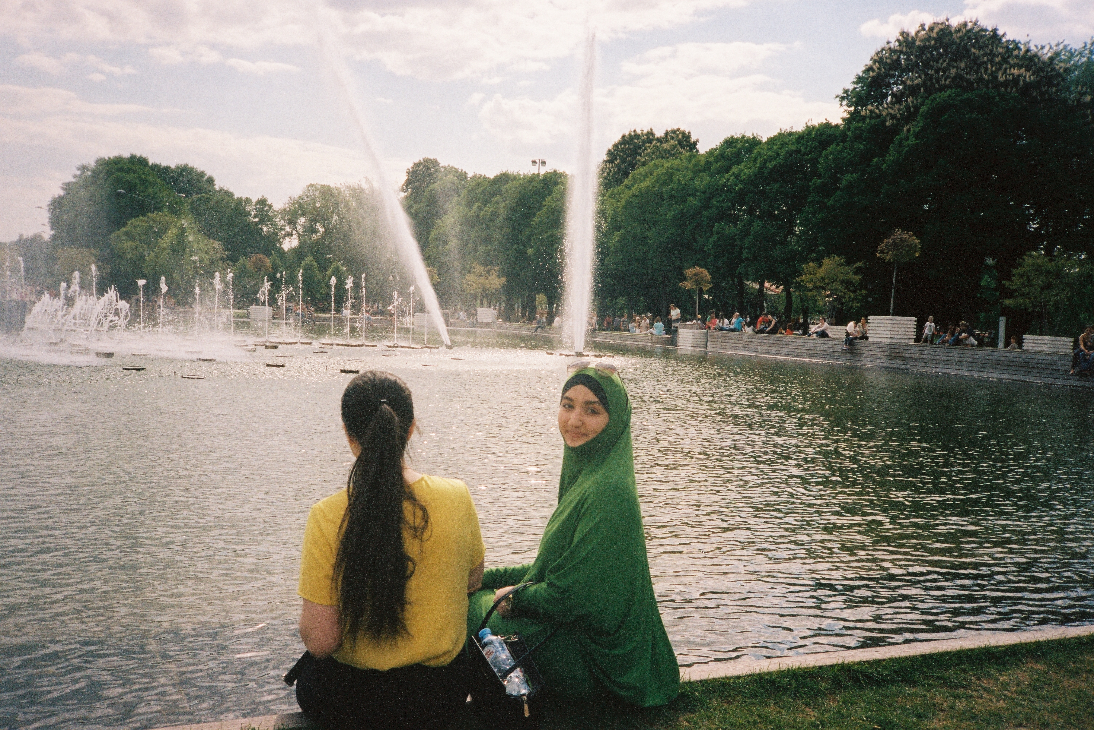 ragazze in riva al lago una con maglietta gialla e coda di cavallo l'altra con hijab color smeraldo
