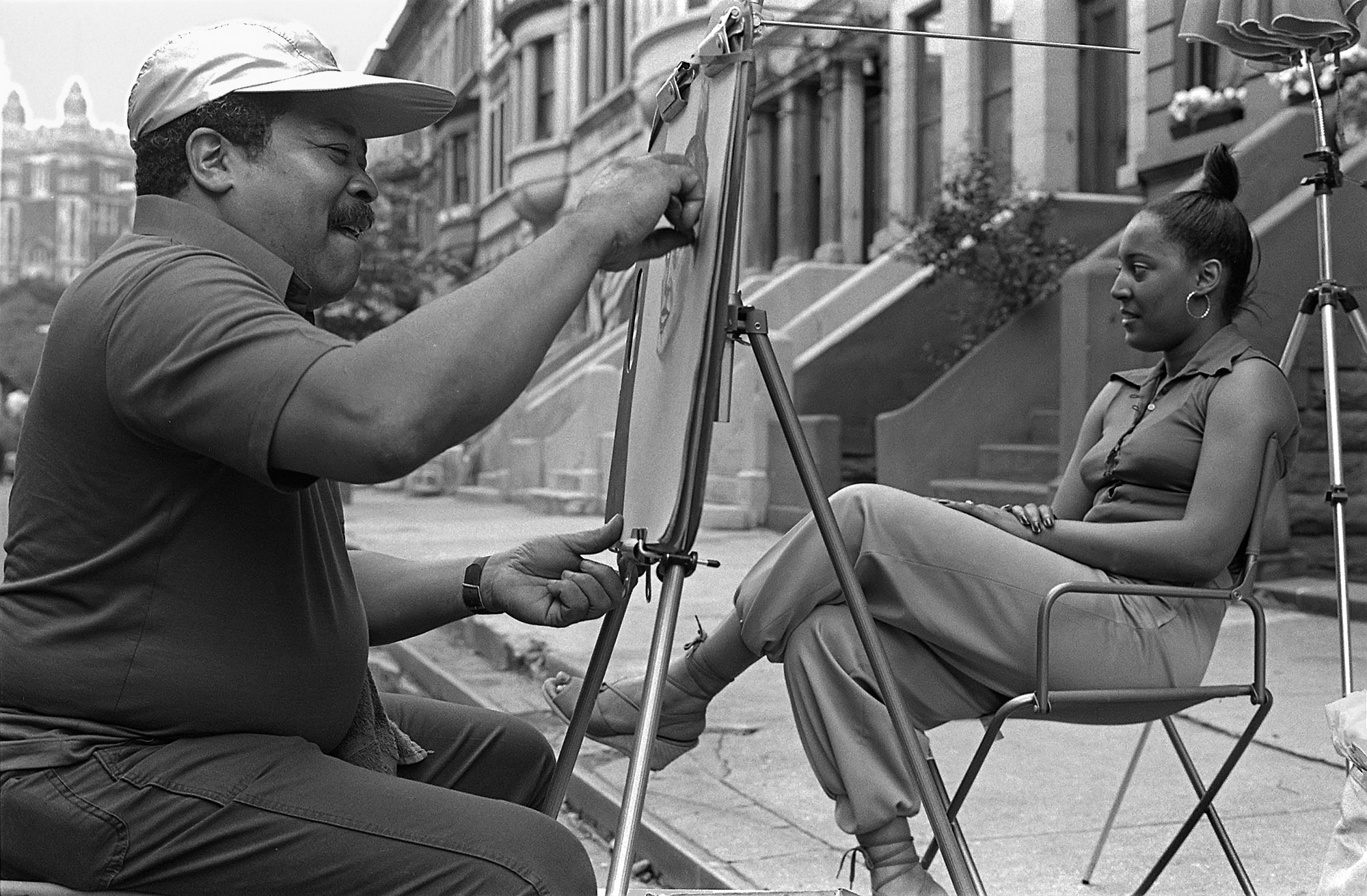 a black and white photo of a street artist drawing a woman sitting in front of him on a chair