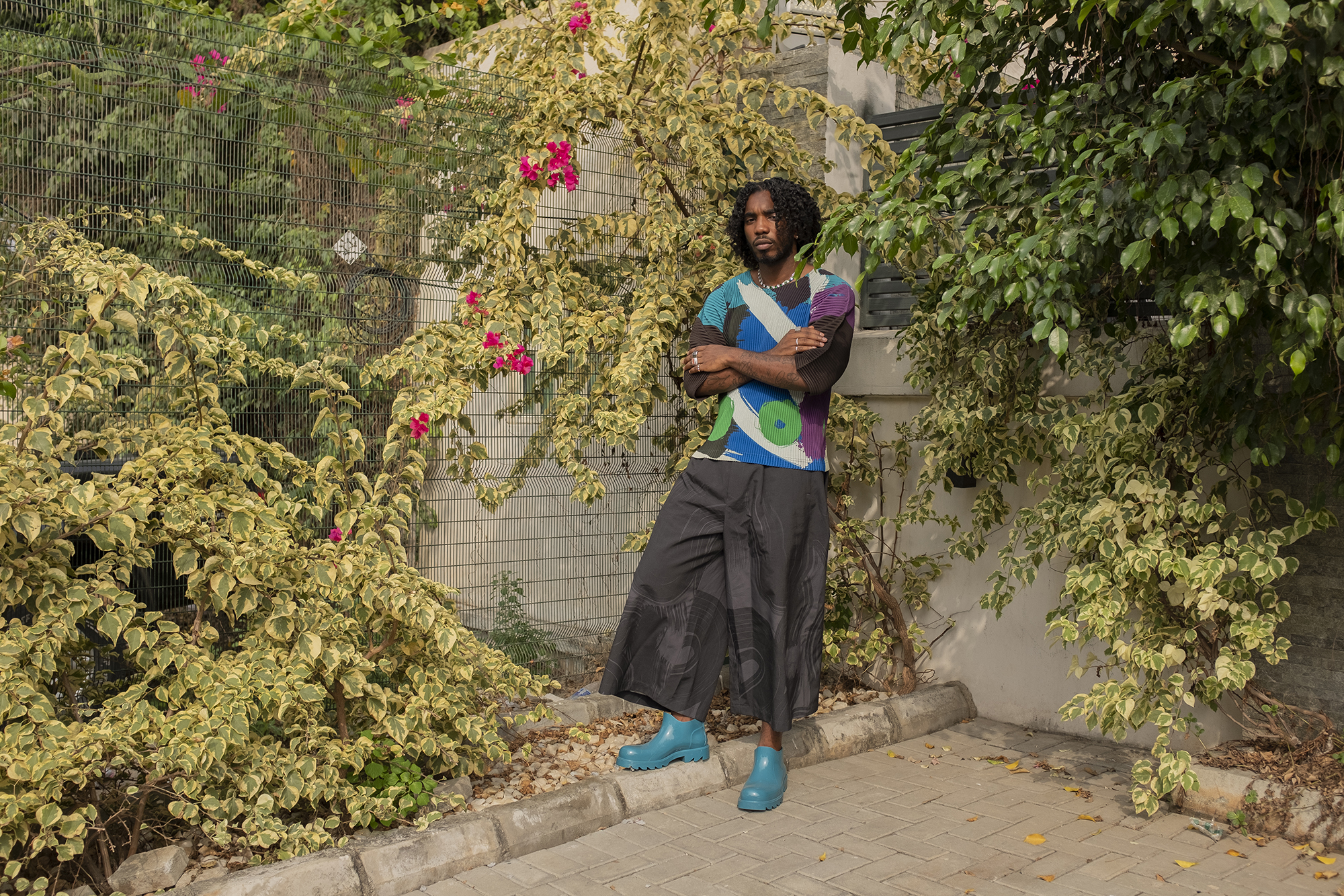 the musician teezee stands in the corner of a garden with his arms folded