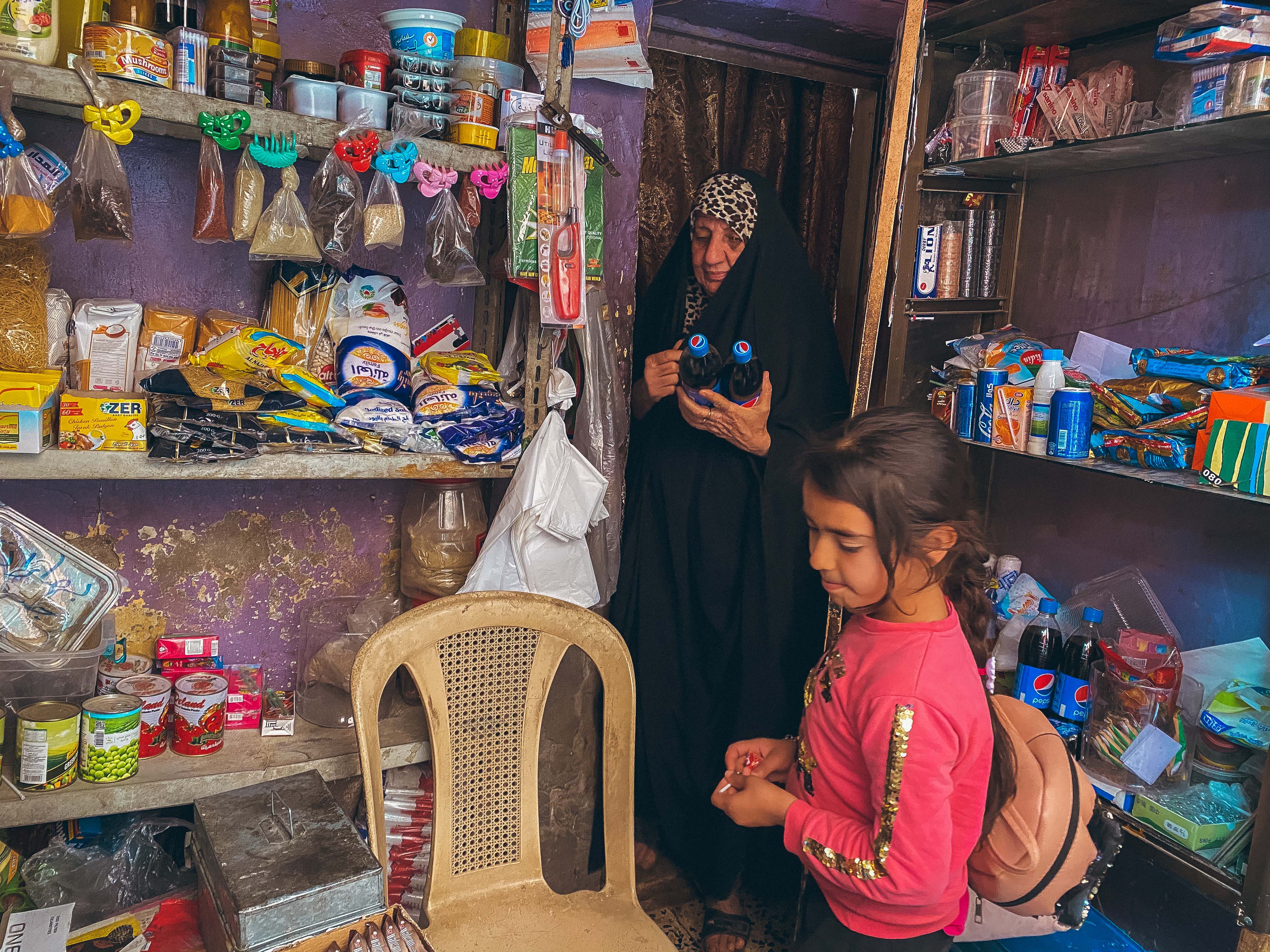 Seorang ibu melayani anak perempuan membeli dua botol pepsi di warungnya. Distrik Kadhimiya, Baghdad. Maret 2021.