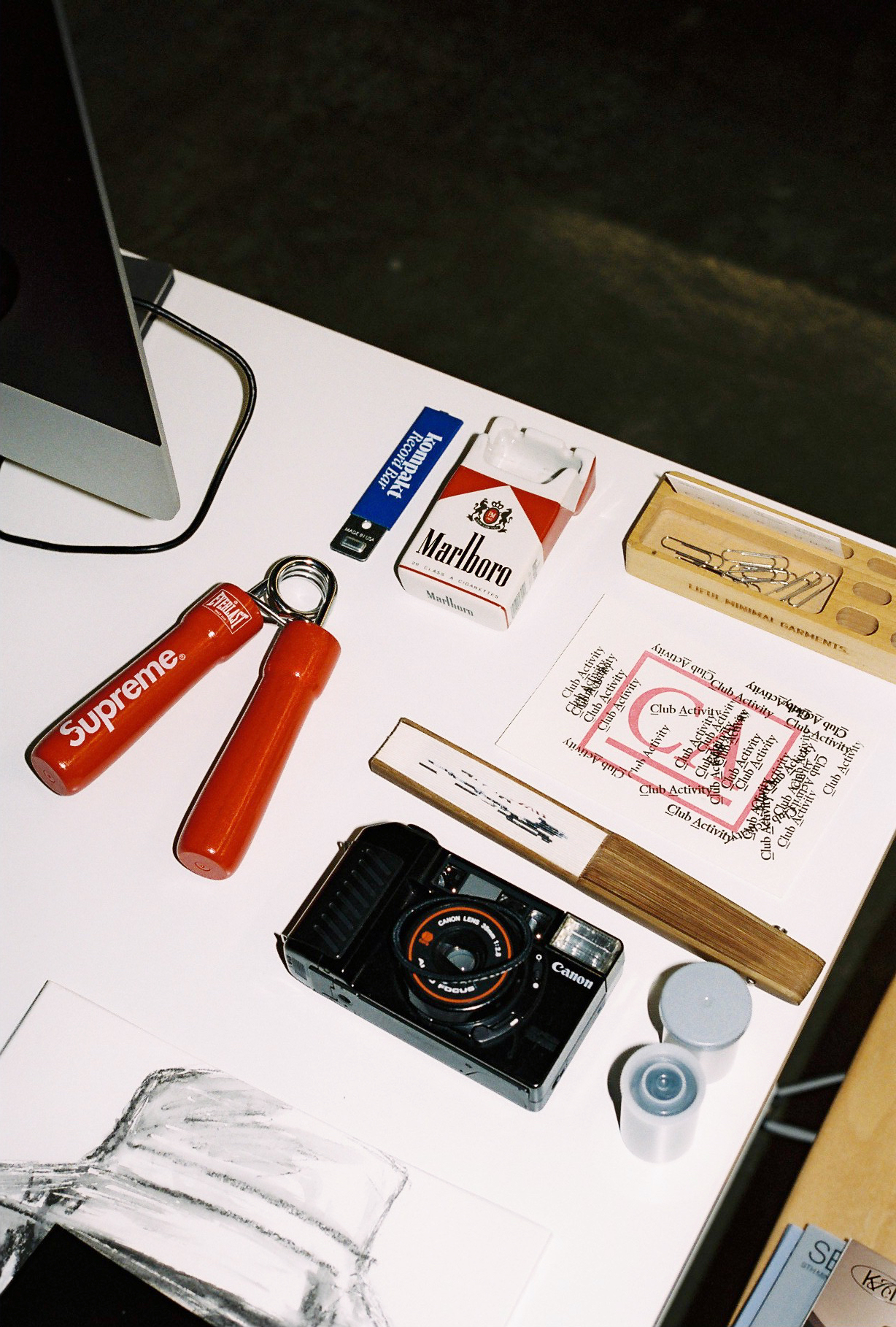 a tidy desk with a camera, supreme knick-knacks, a wooden fan and a marlboro box on it