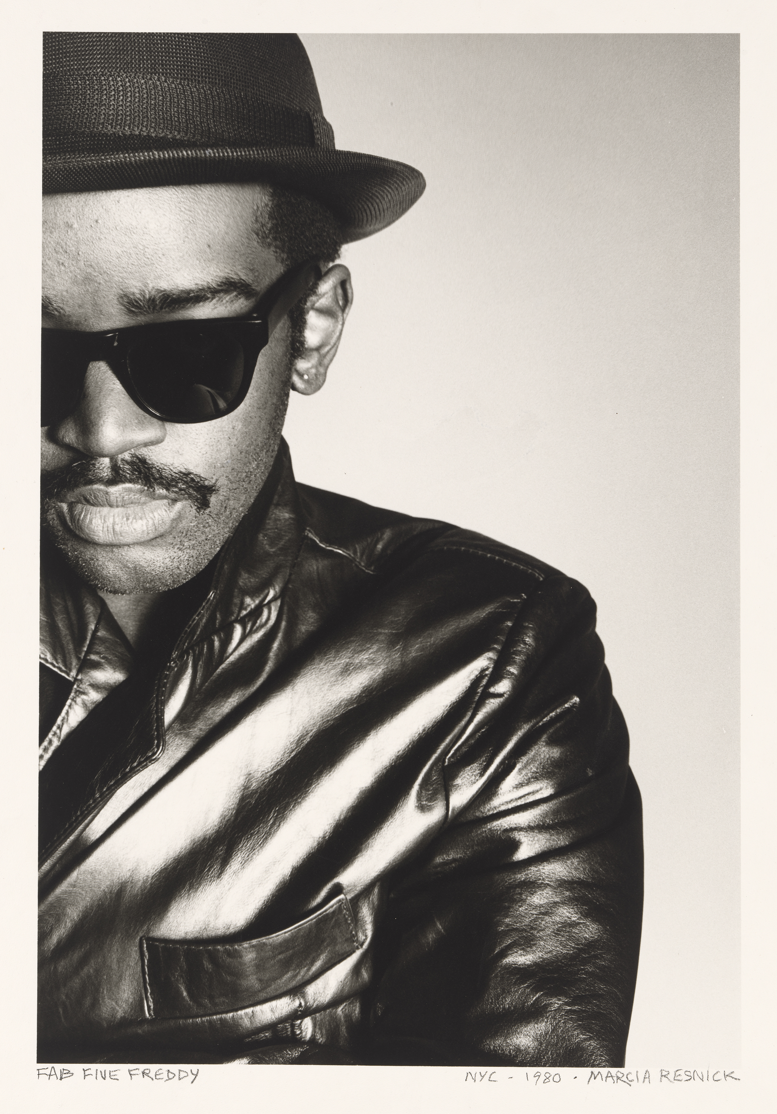 Portrait of Fab 5 Freddy wearing a leather jacket, bowler hat and sunglasses photographed by Marcia Resnick.
