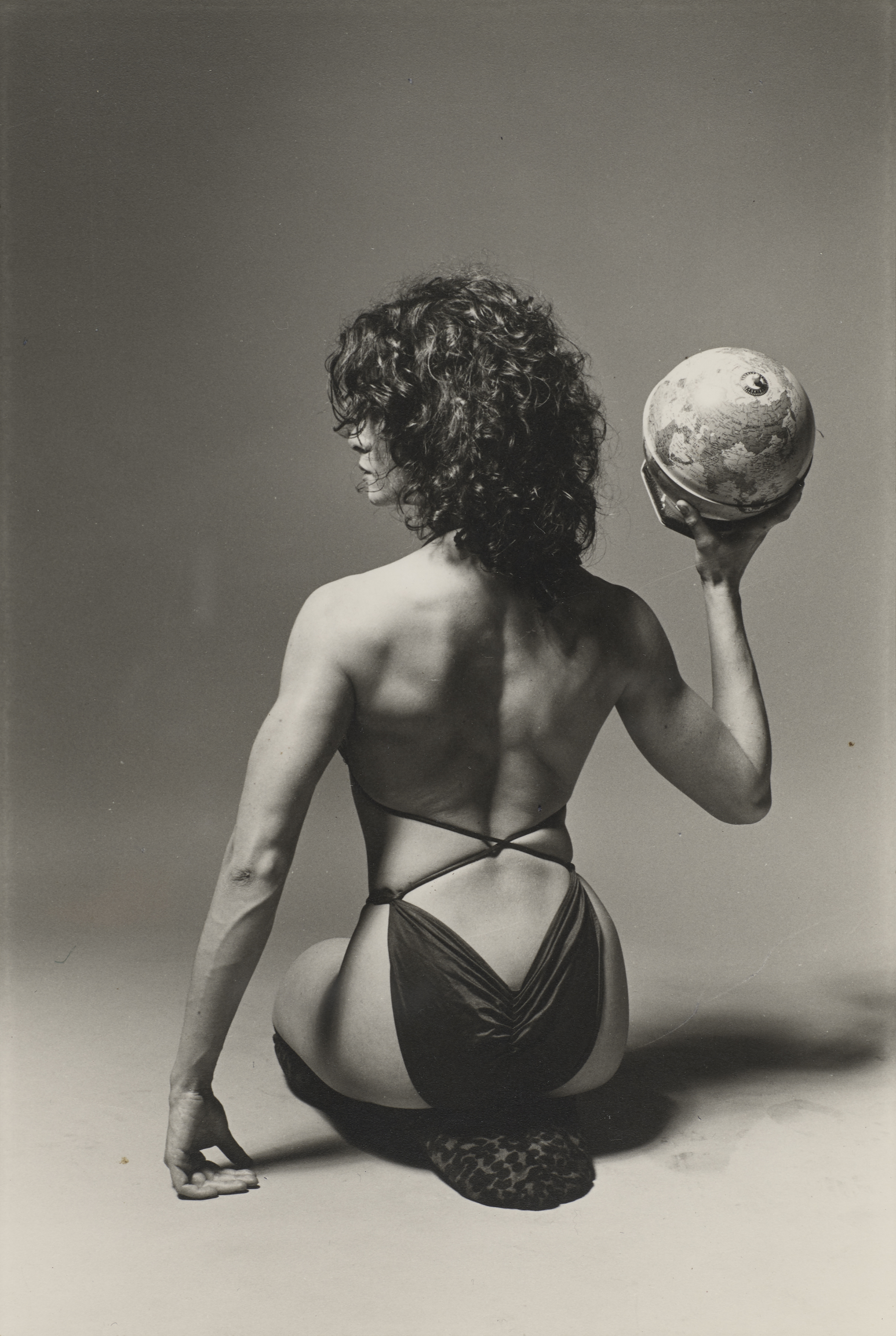 Photo of Lisa Lyon in a bikini and holding a ball and sitting with her back to the camera, photographed by Marcia Resnick.