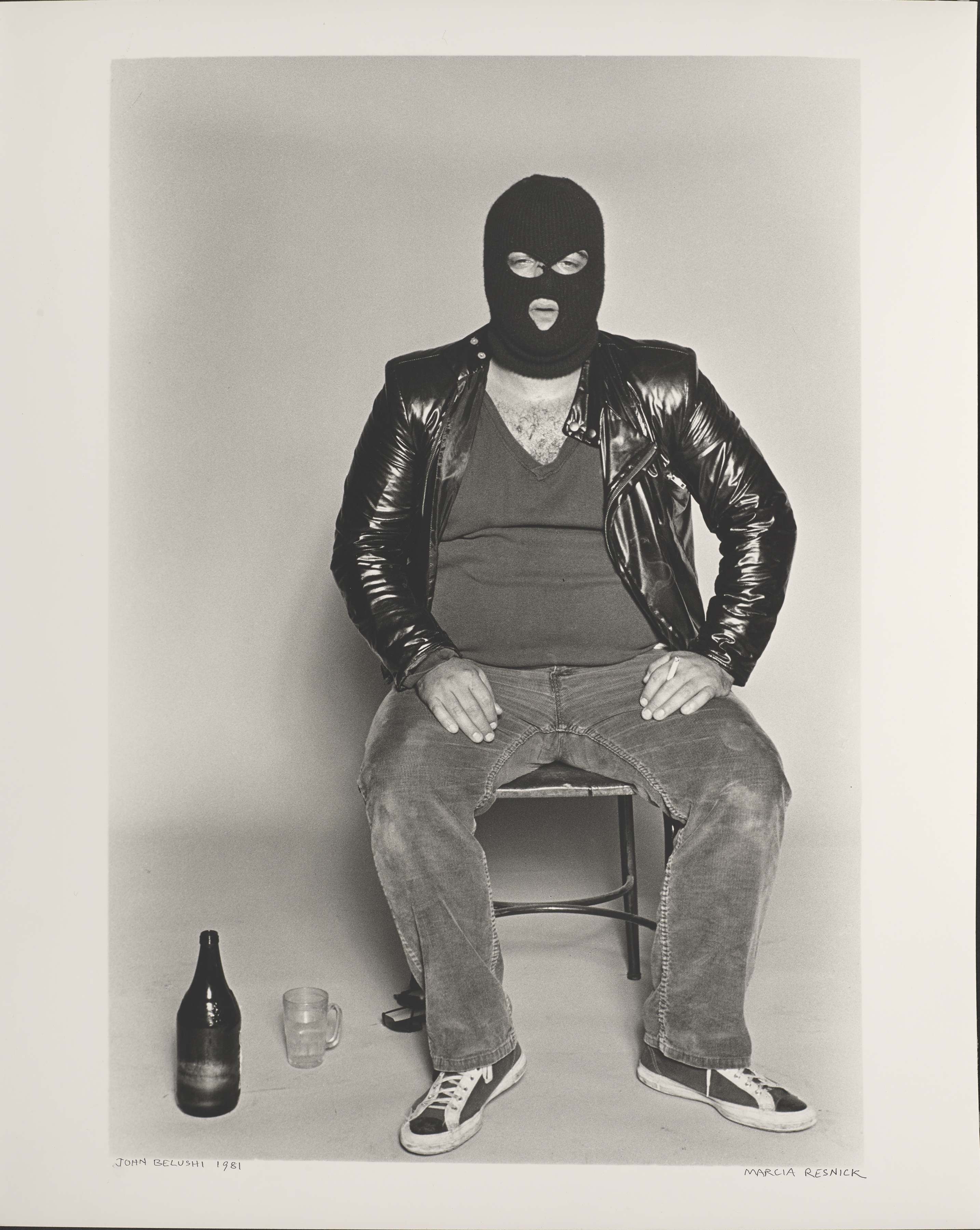 Portrait of John Belushi sitting down on a chair in a vest, jeans, trainers, latex jacket and face mask with a drink and glass next to him photographed by Marcia Resnick.