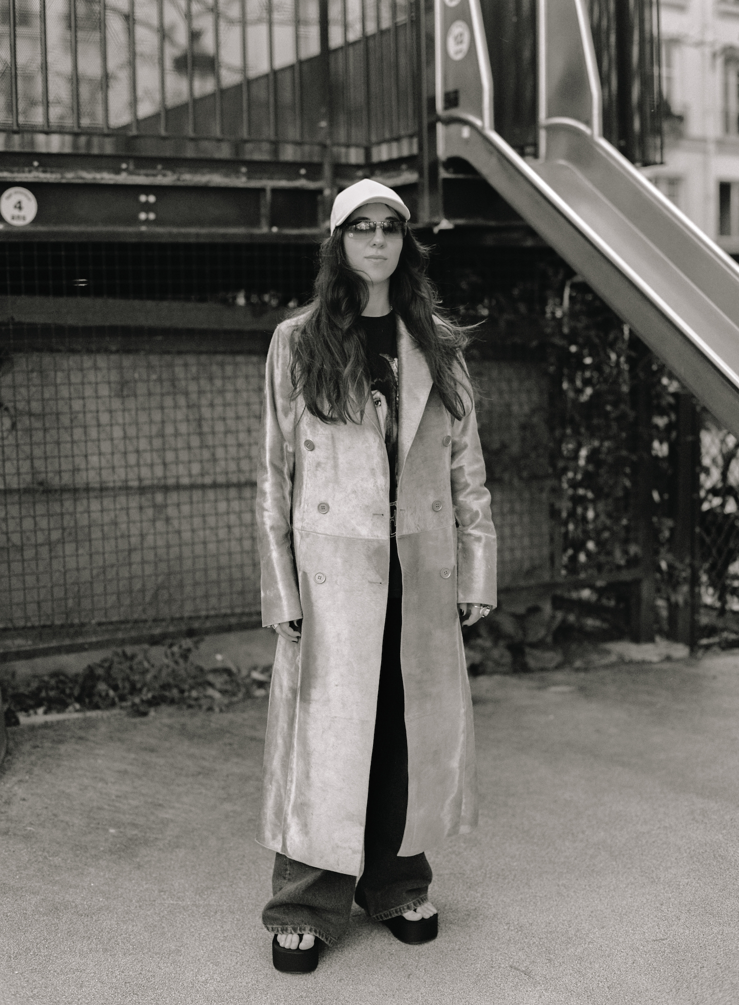 Chloe Caillet wearing a ling leather coat, cap and sunglasses, standing by a slide 