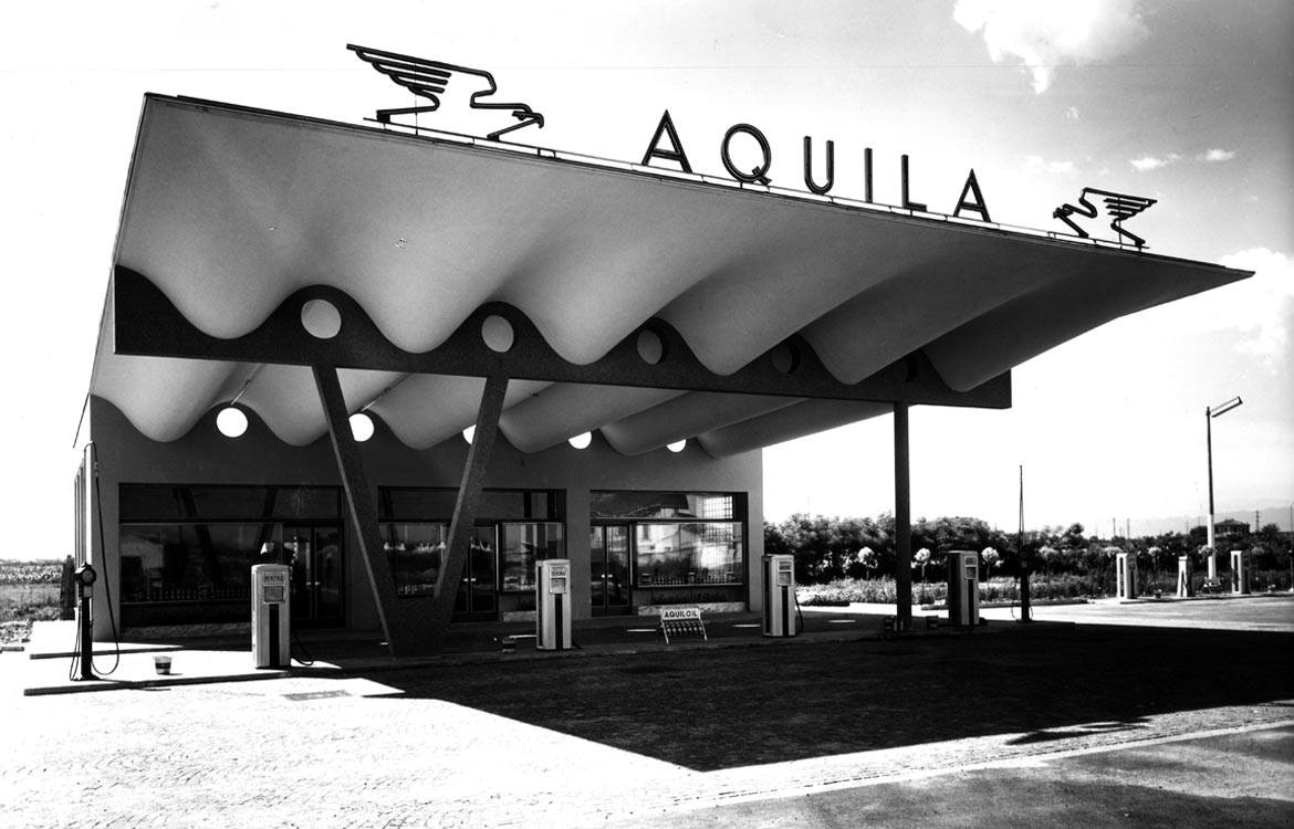 Forgotten Architectures, Aquila gas station,  Aldo Favini – black and white photo of a gas station with a roof supported by beams with a curved pattern with round cutouts. On top of it, there's a sign reading 