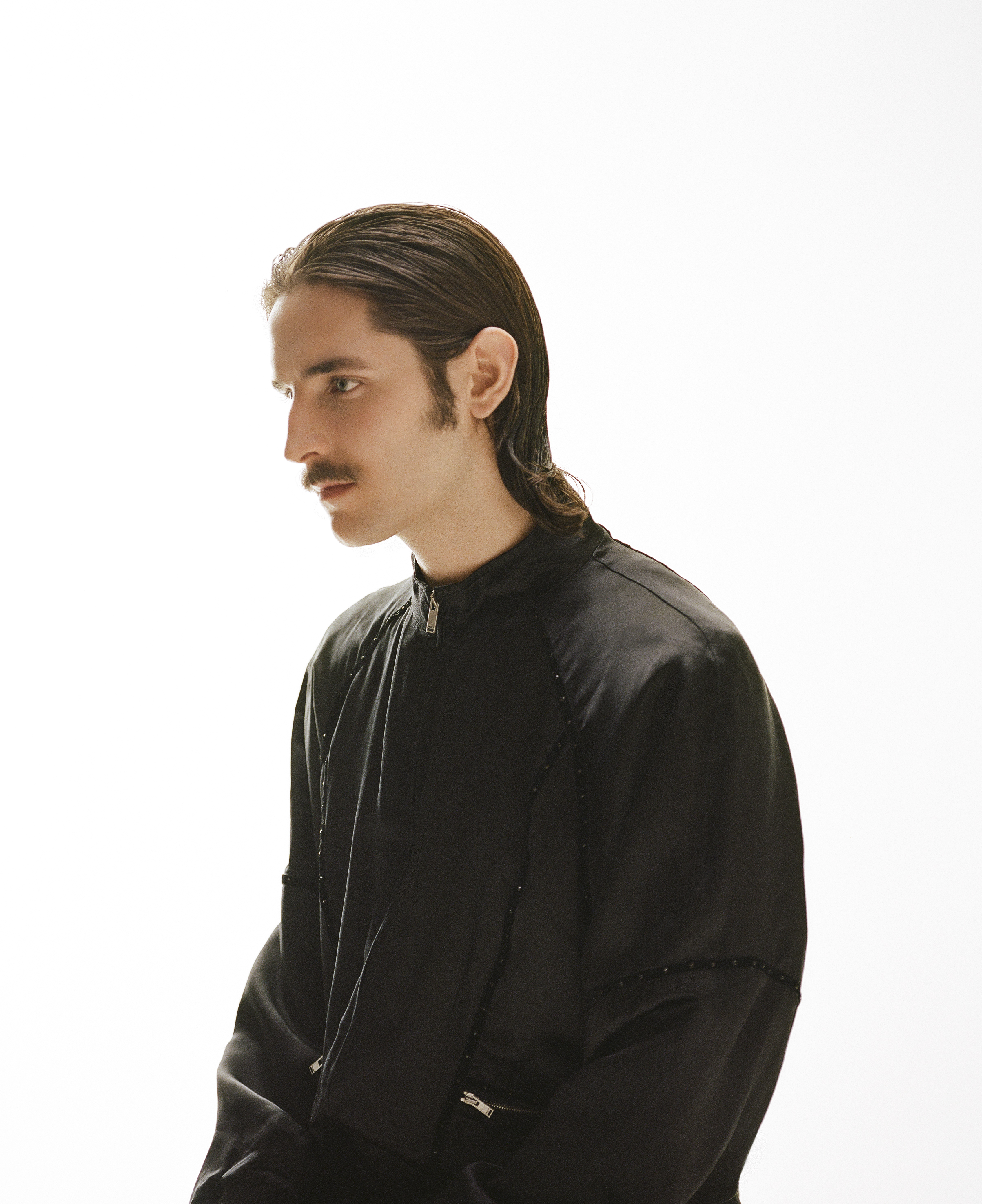 a member of the band Keep Dancing Inc, in a black jacket with slicked back hair and a moustache
