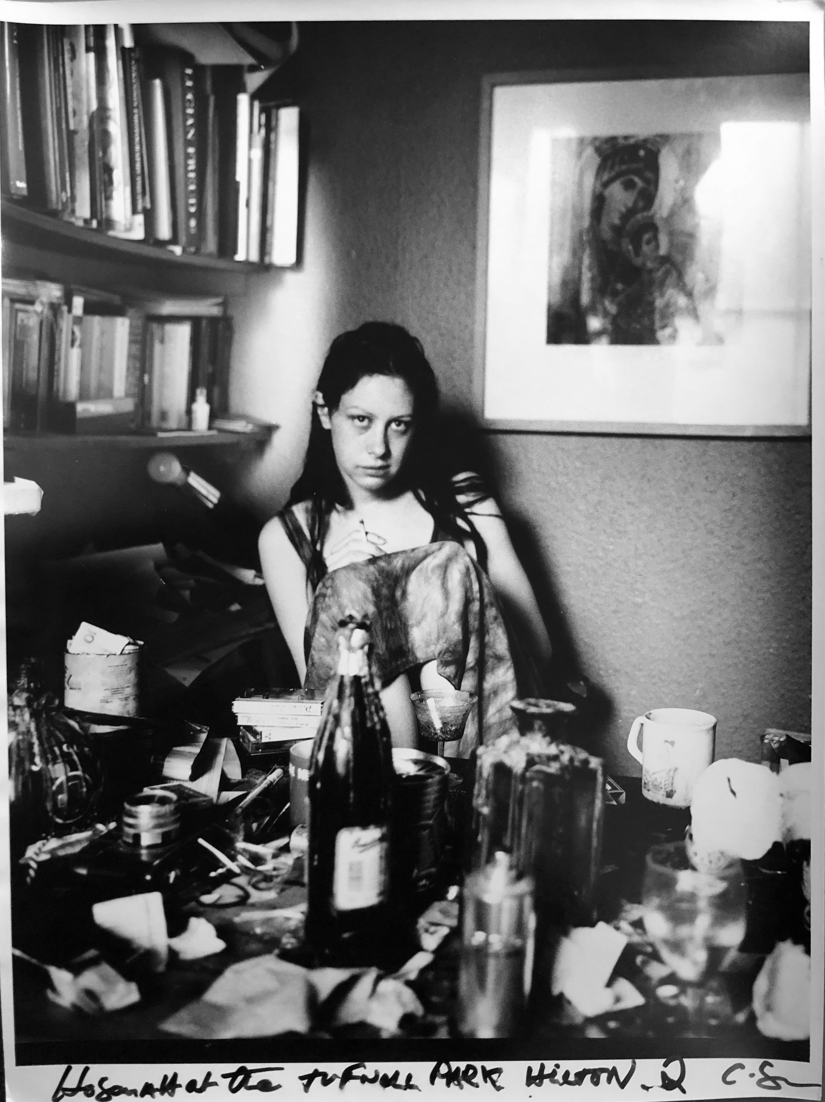 black-and-white image of a woman sitting at a table littered with bottles, glasses and cameras