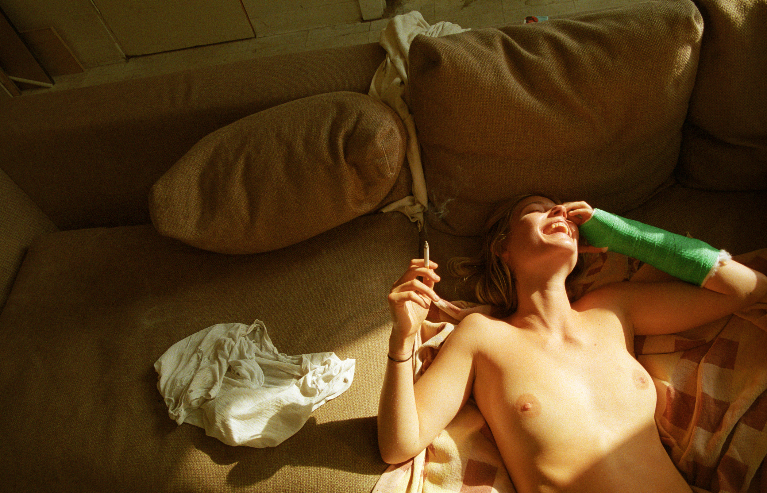 a person with a green cast on their arm laughs as they smoke a cigarette, laying topless on their sofa