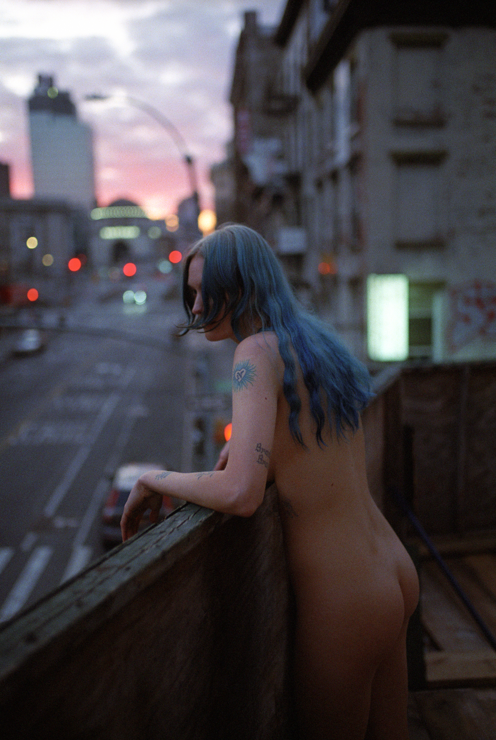 a person with long blue hair stands nude on a city rooftop 