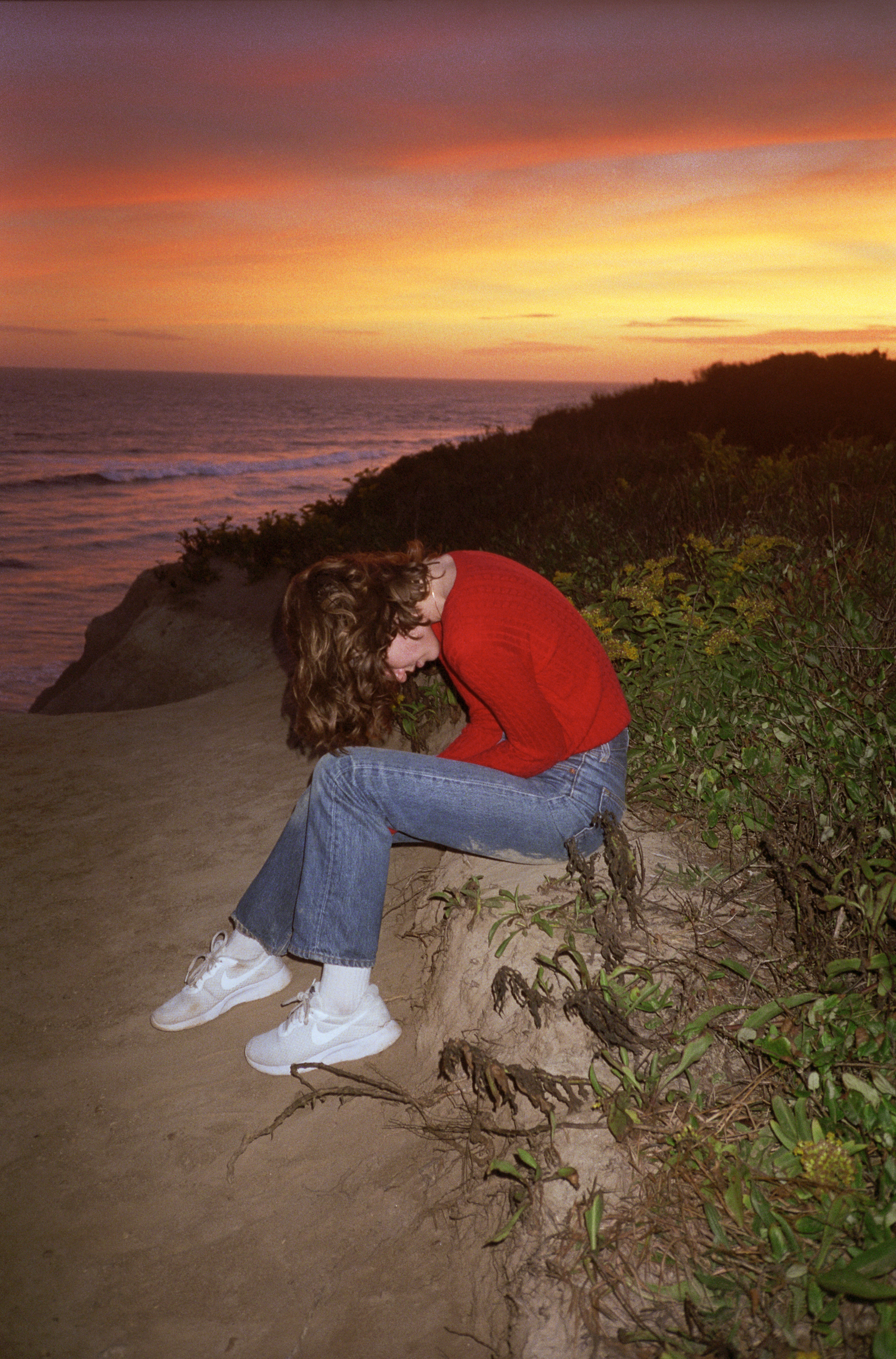a person in a red sweater and jeans leans over in front of a beach sunset