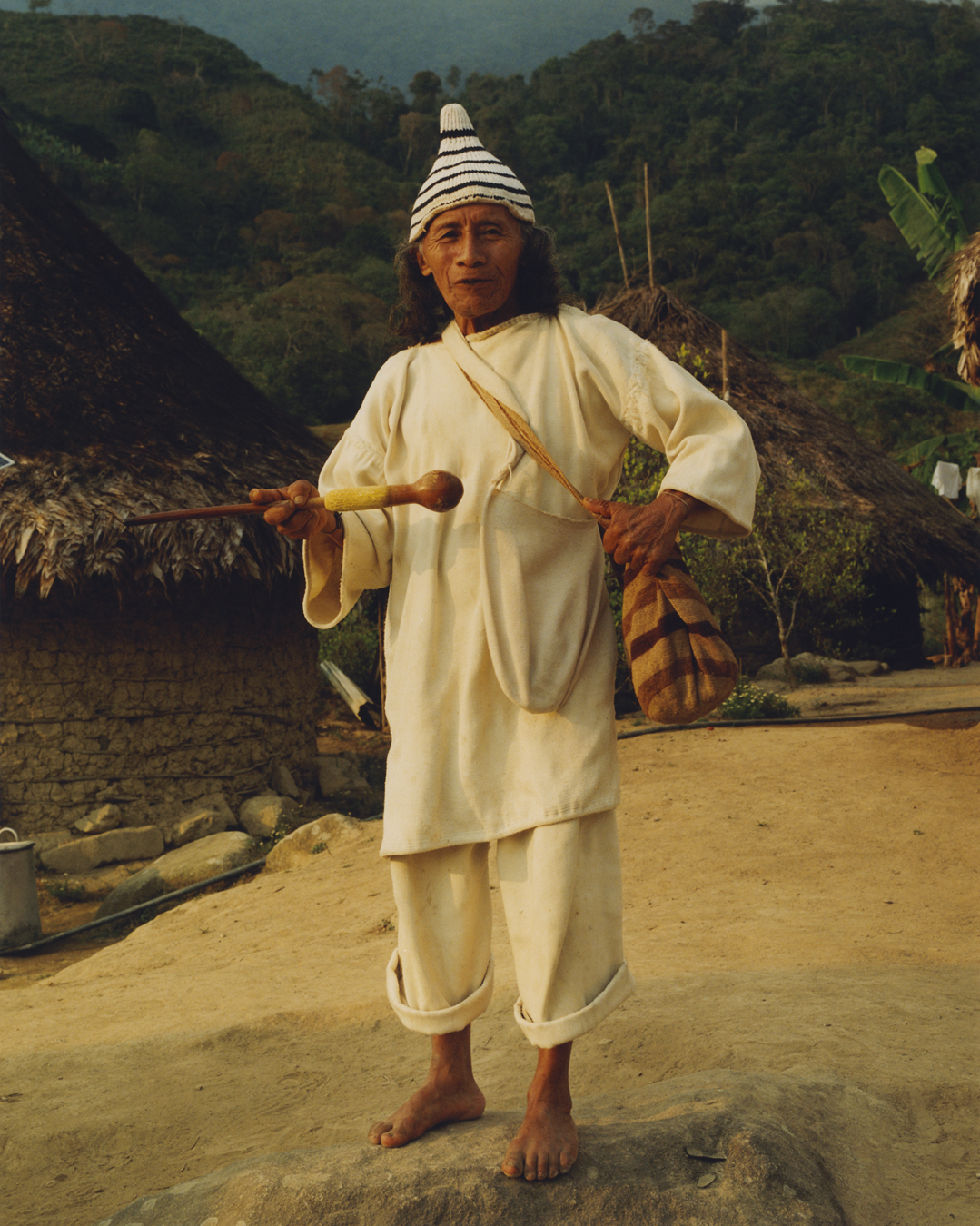 Man in white cotton clothing with a brown bag, a wooden stick and a stripy hat.