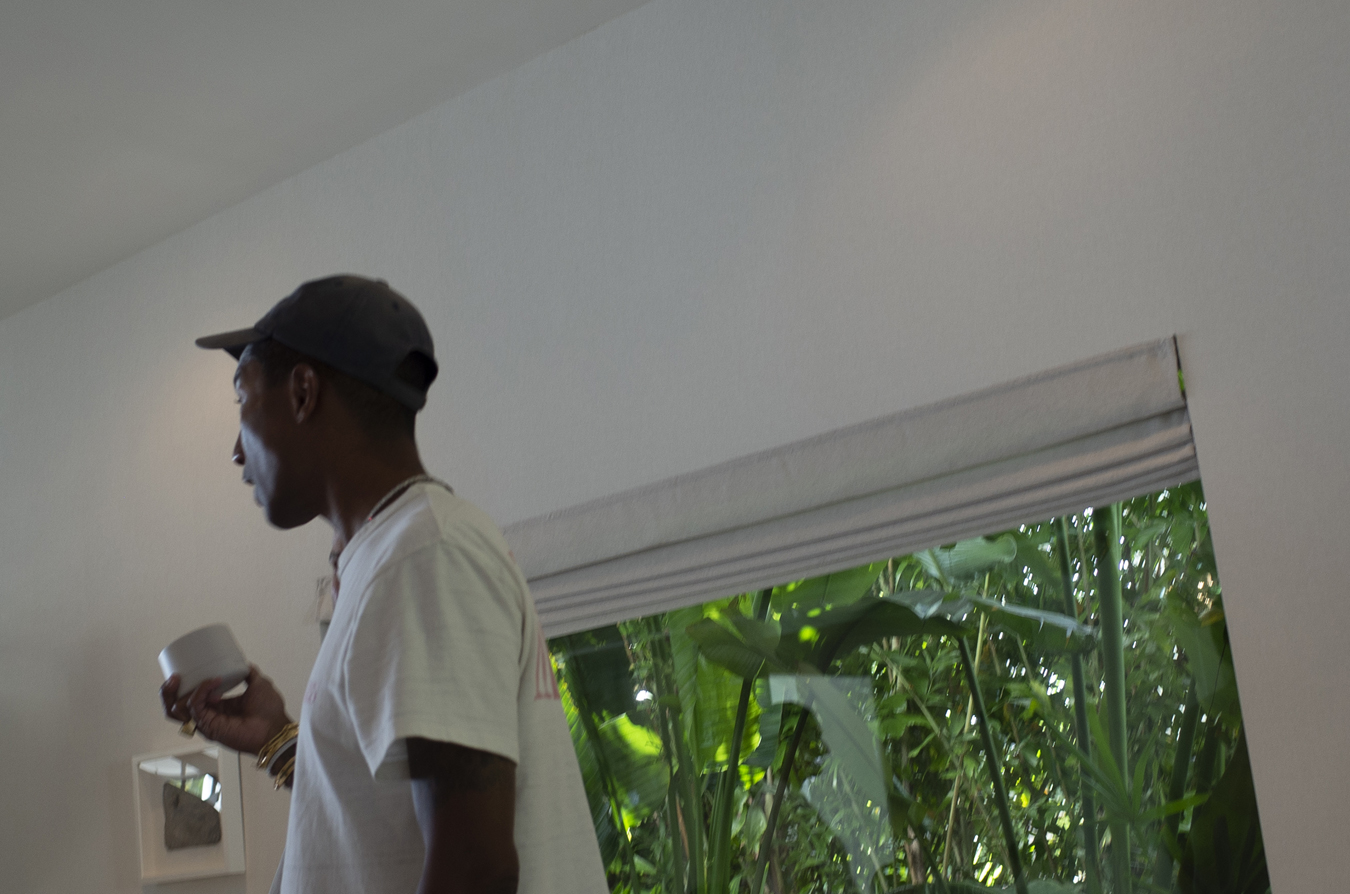Pharrell Williams sosteniendo una crema para la piel Humanrace y de pie junto a la ventana.