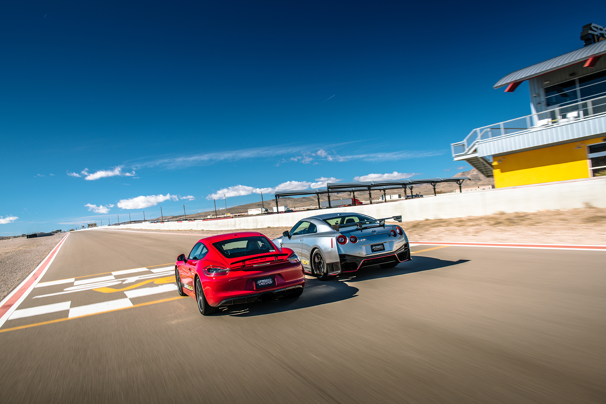 Two exotic sportscars racing next to each other on a track beneath a blue sky. 
