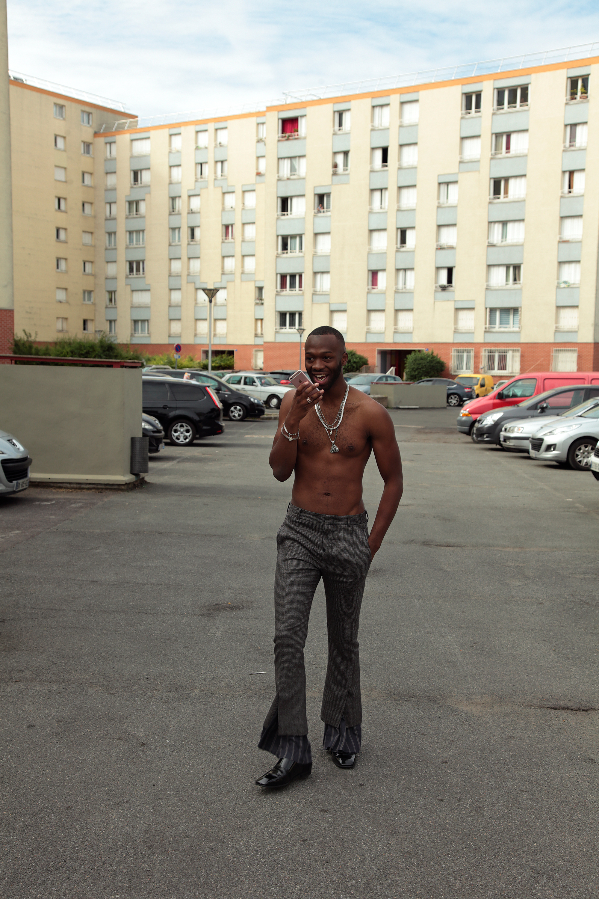 a topless man in grey bootcut pants walks aross a car park talking on a pink iphone 