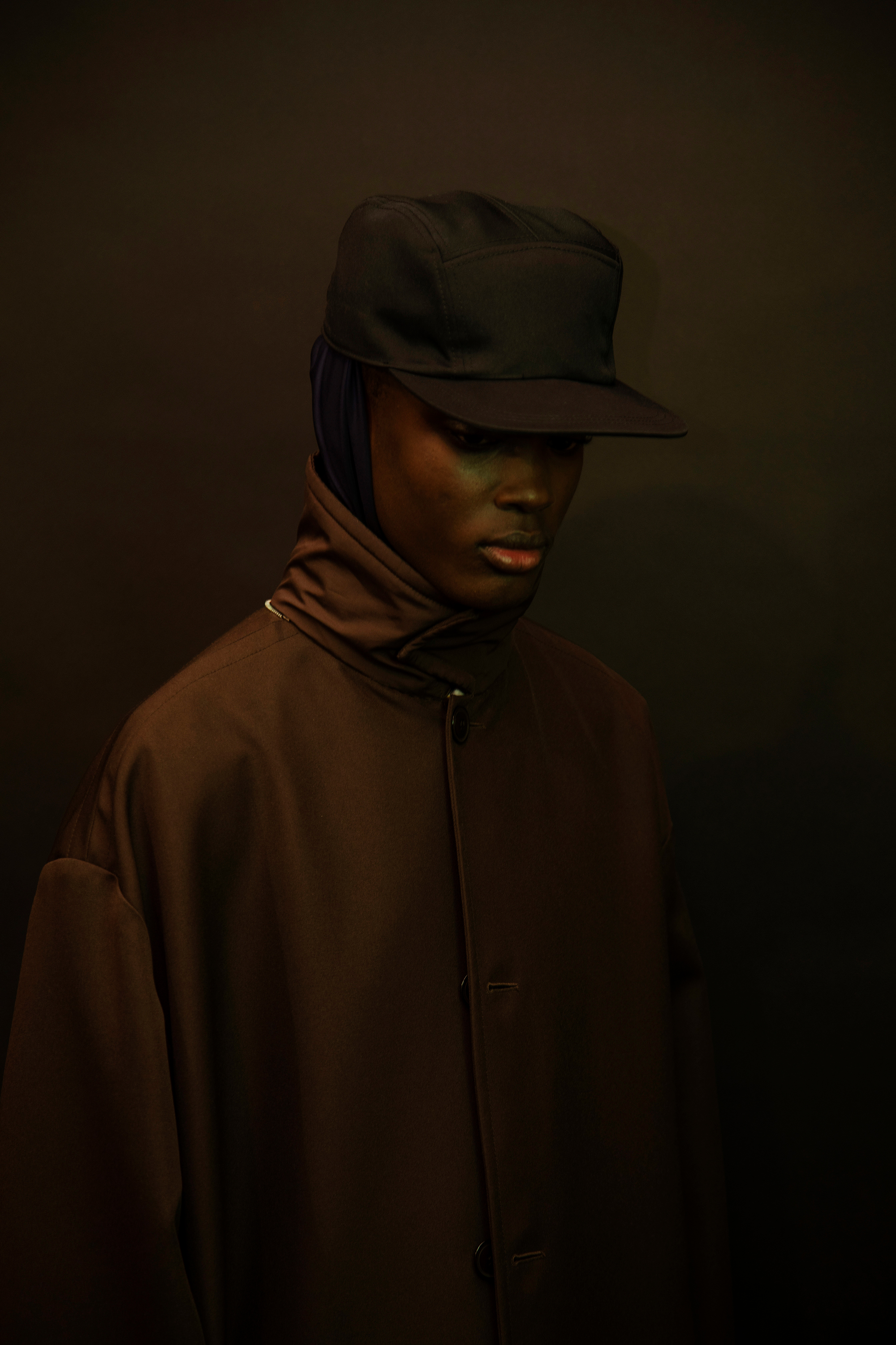 a man in a brown coat and baseball cap shot in low light semi-profile 