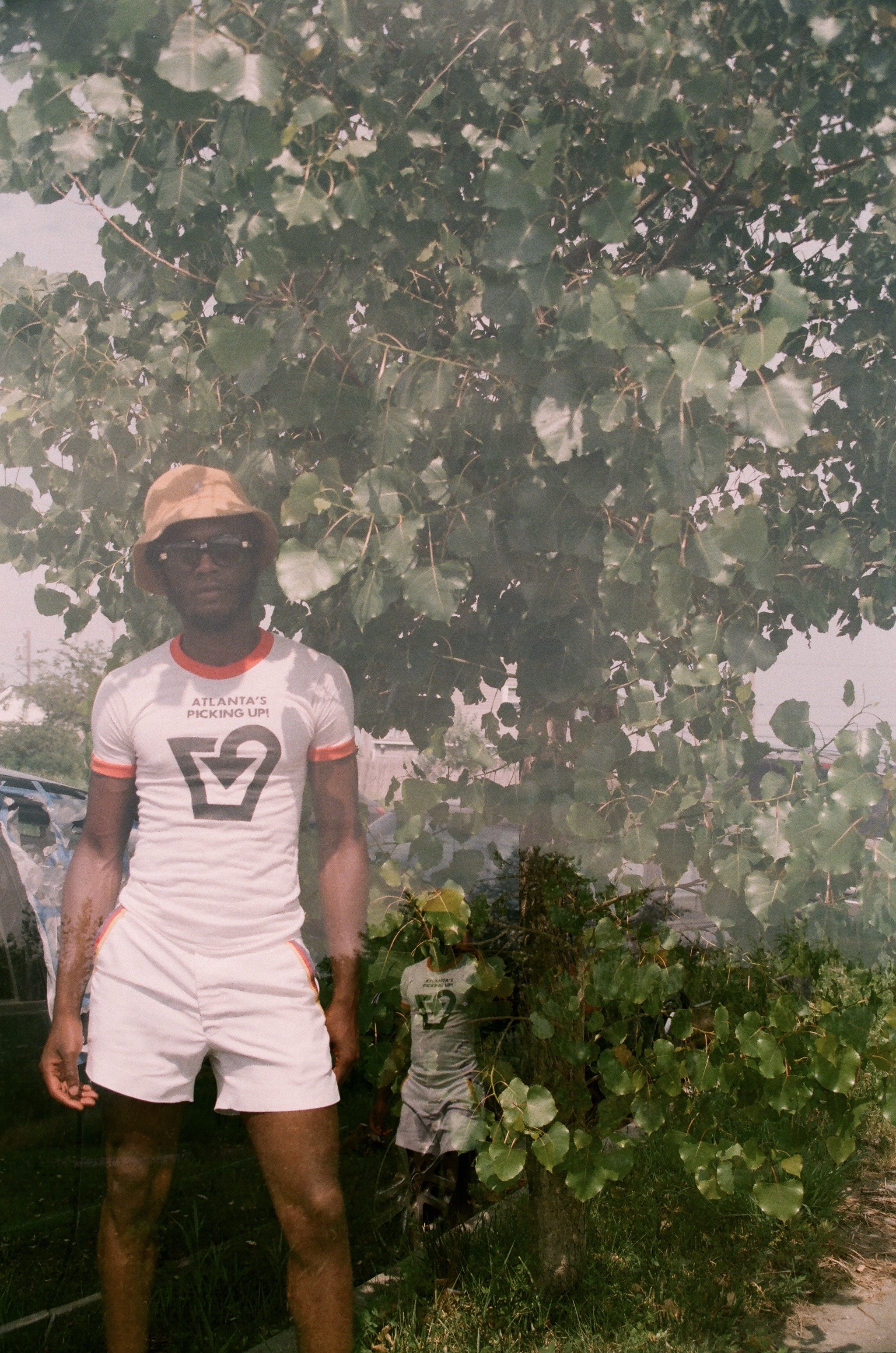A man in white shorts and t-shirt standing in front of a bush in Queens