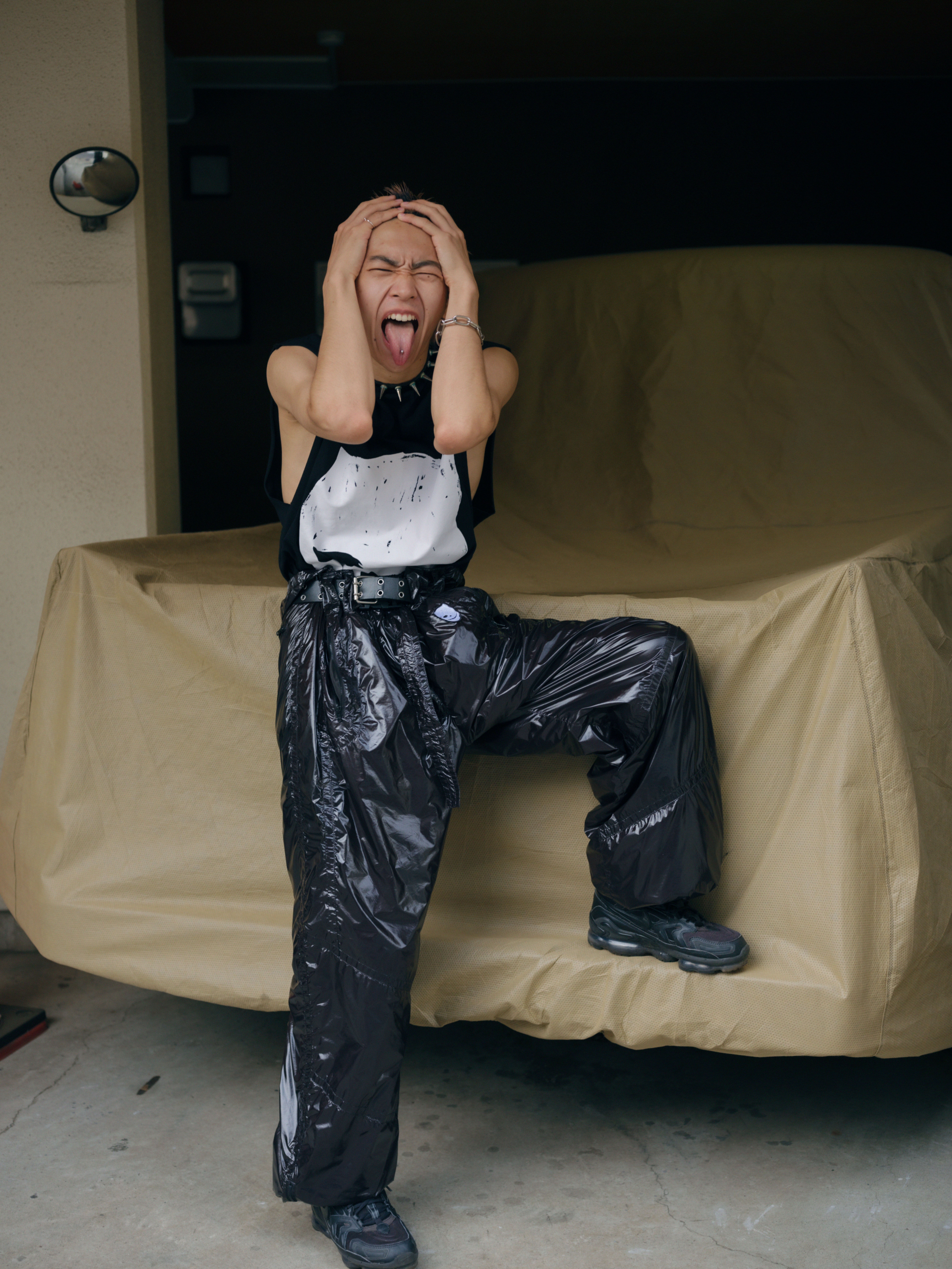 a person wearing shiny parachute trousers sticks their pierced tongue out to camera, with their head in their hands